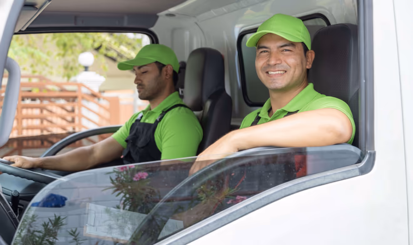 Two delivery men in green uniforms inside a white truck, one smiling and leaning out the window, the other focused on driving.