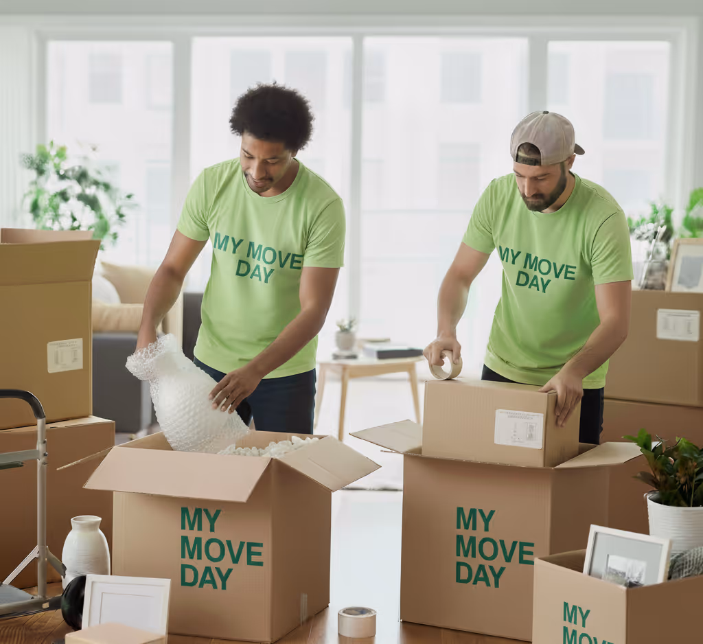 Two men in green 'My Move Day' t-shirts packing household items into cardboard boxes inside a bright room.