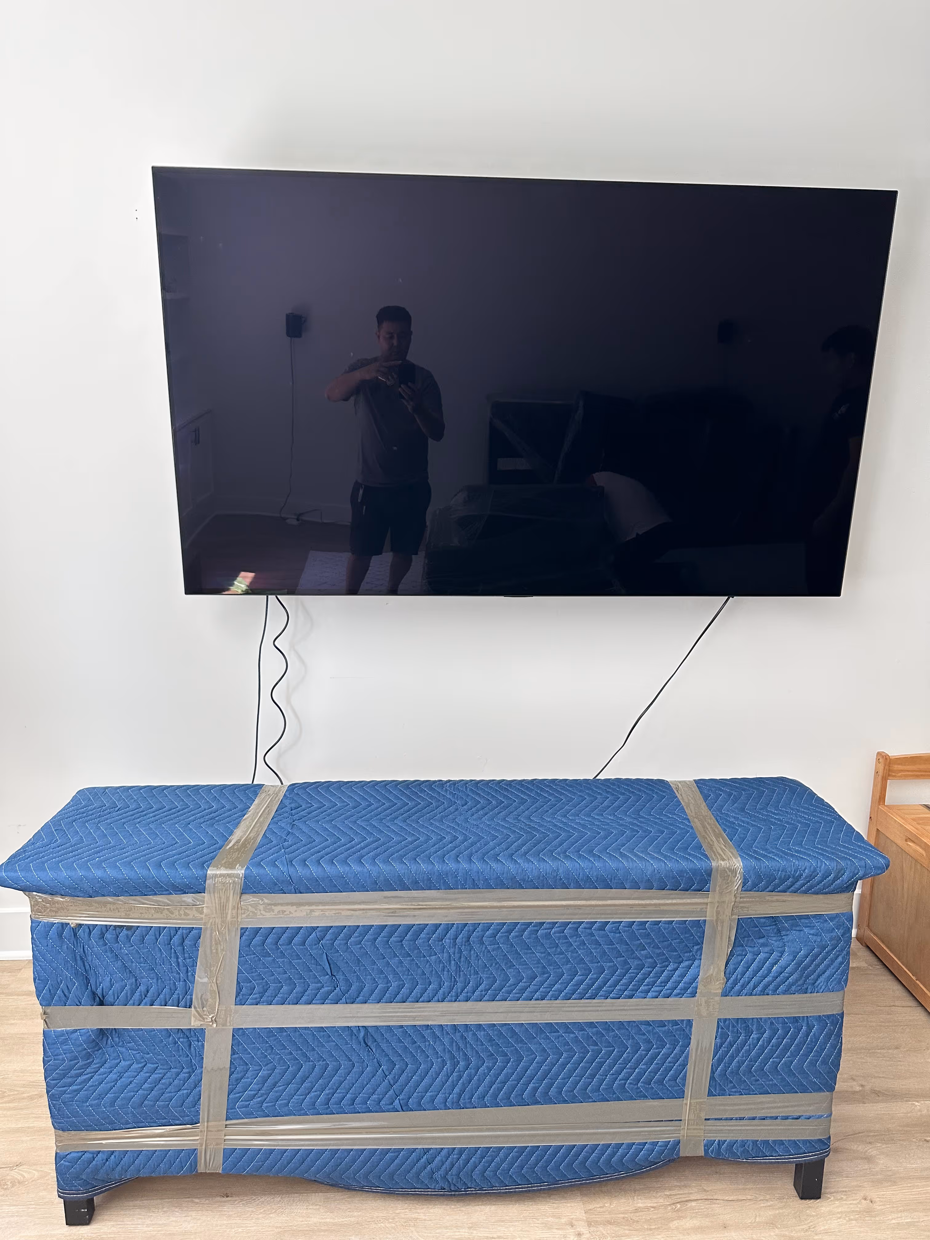 Flat-screen TV mounted on wall above a blue quilted moving blanket wrapped and taped around a piece of furniture in a room with light wood flooring.