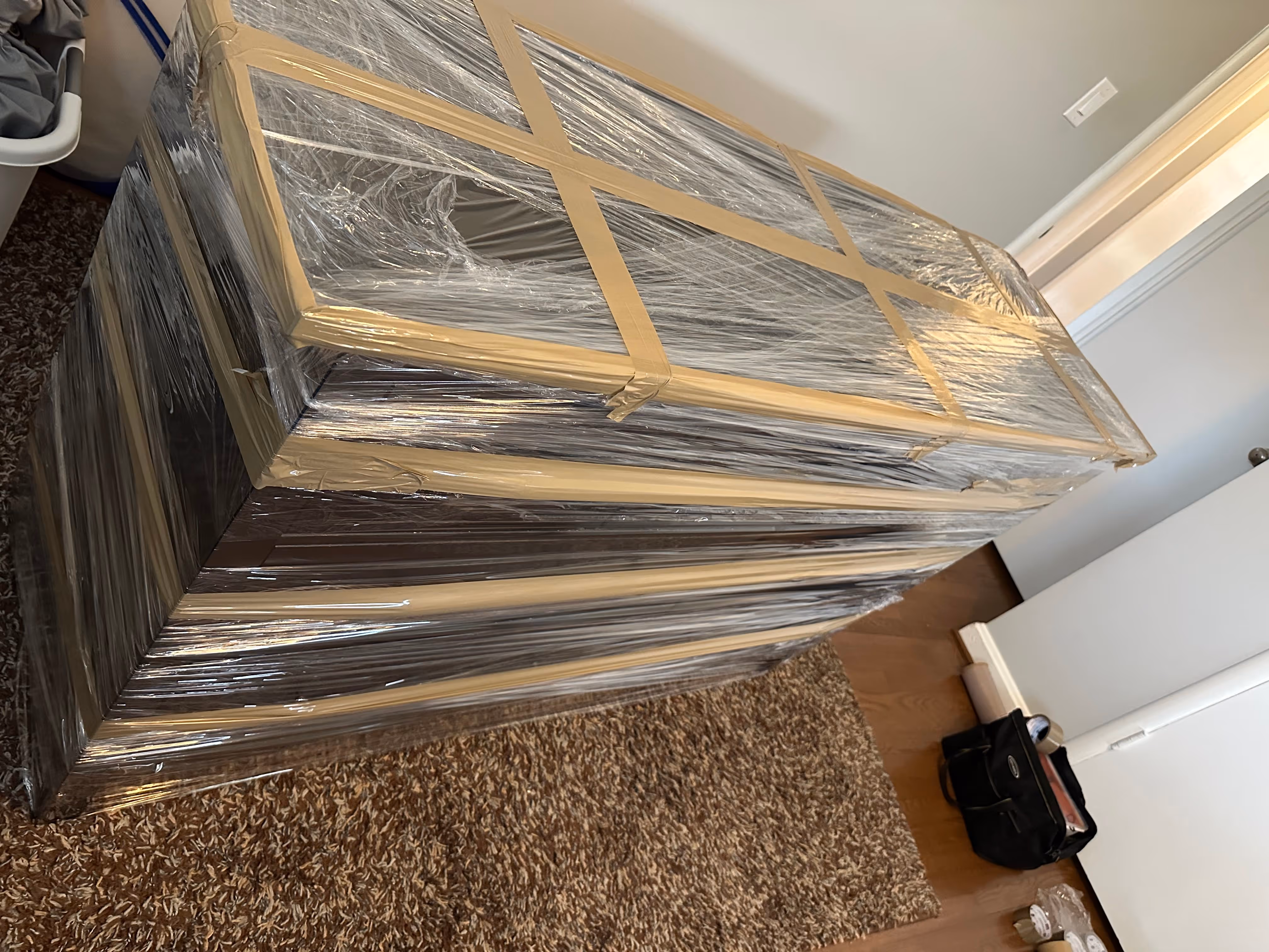 Tall cabinet wrapped in clear plastic and beige tape on a brown carpet in a room with wooden floor.