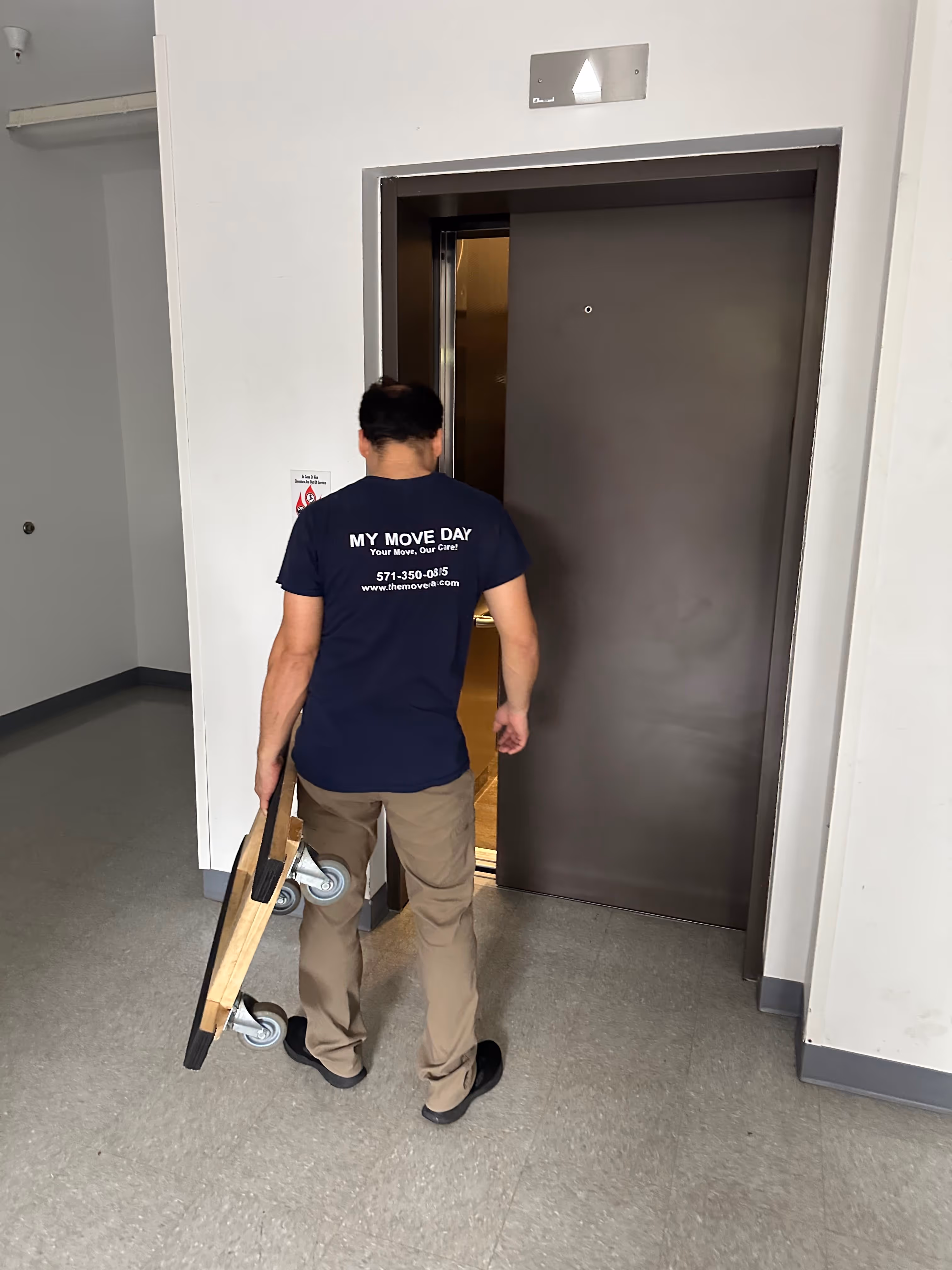 Person wearing a 'My Move Day' shirt carrying a dolly, walking toward an open elevator door in a plain hallway.