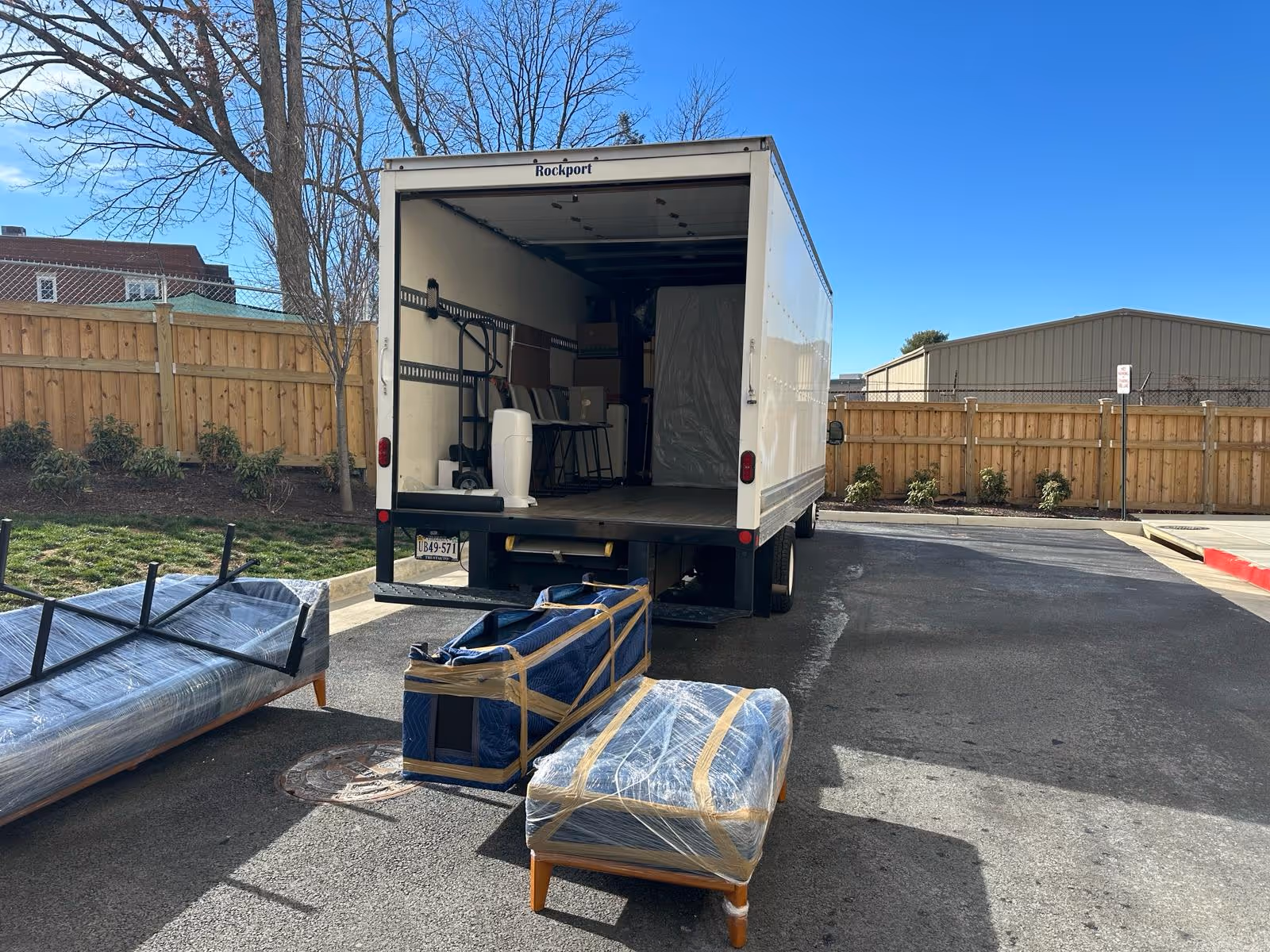 White moving truck with open back doors parked near a wooden fence, surrounded by wrapped and taped furniture pieces on asphalt under a clear blue sky.