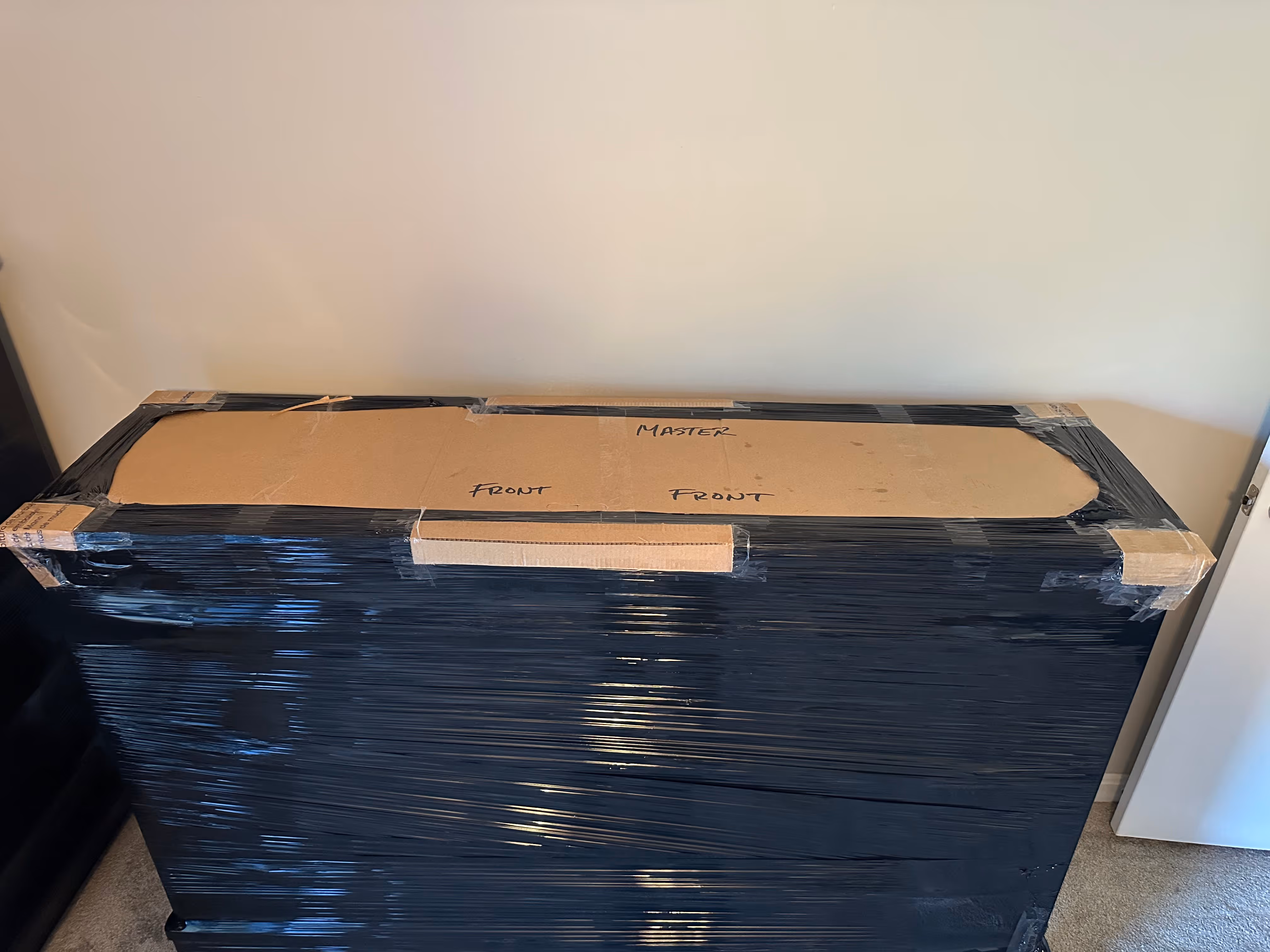 Large rectangular item wrapped in black plastic wrap with cardboard taped on top labeled 'MASTER' and 'FRONT'.