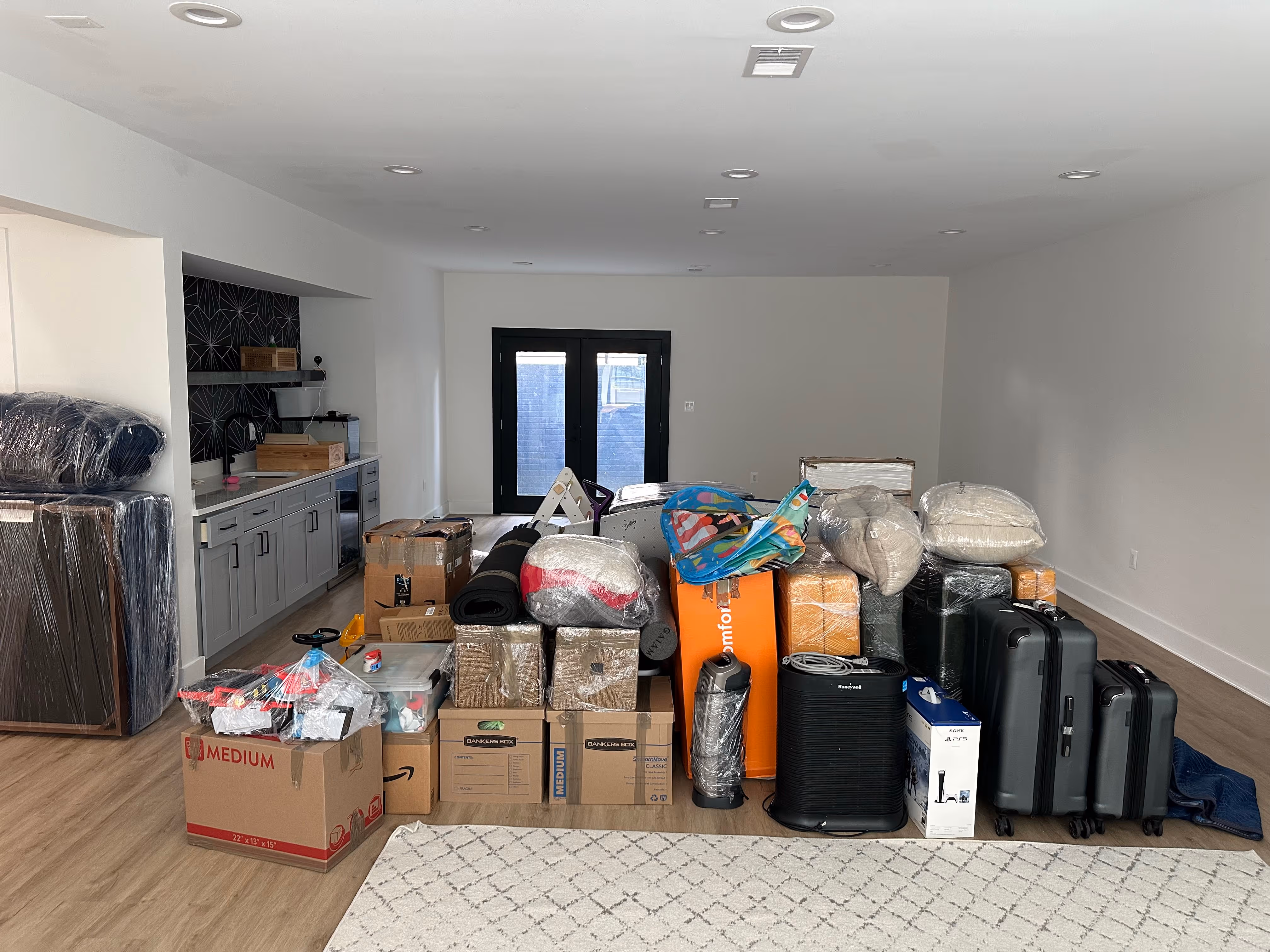Stacked moving boxes, wrapped furniture, suitcases, and household items in a spacious room with wooden floors and white walls.