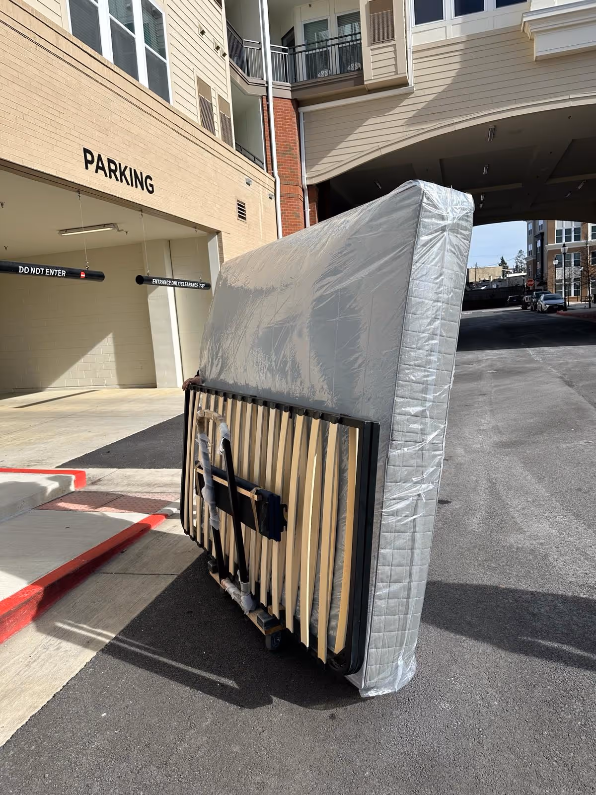 Large mattress wrapped in plastic and a bed frame leaning upright on an asphalt street near a building with parking signs.