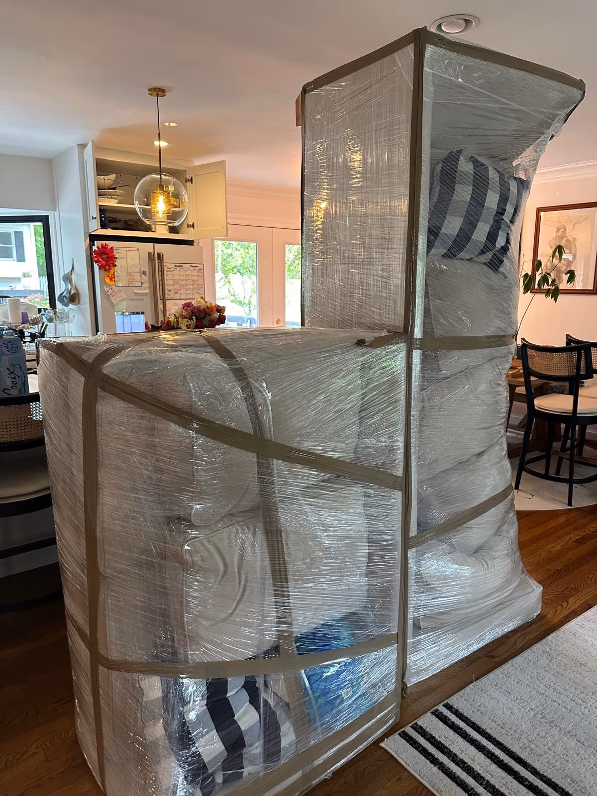 Two large furniture pieces wrapped in plastic and tape inside a living space with kitchen and dining area visible.