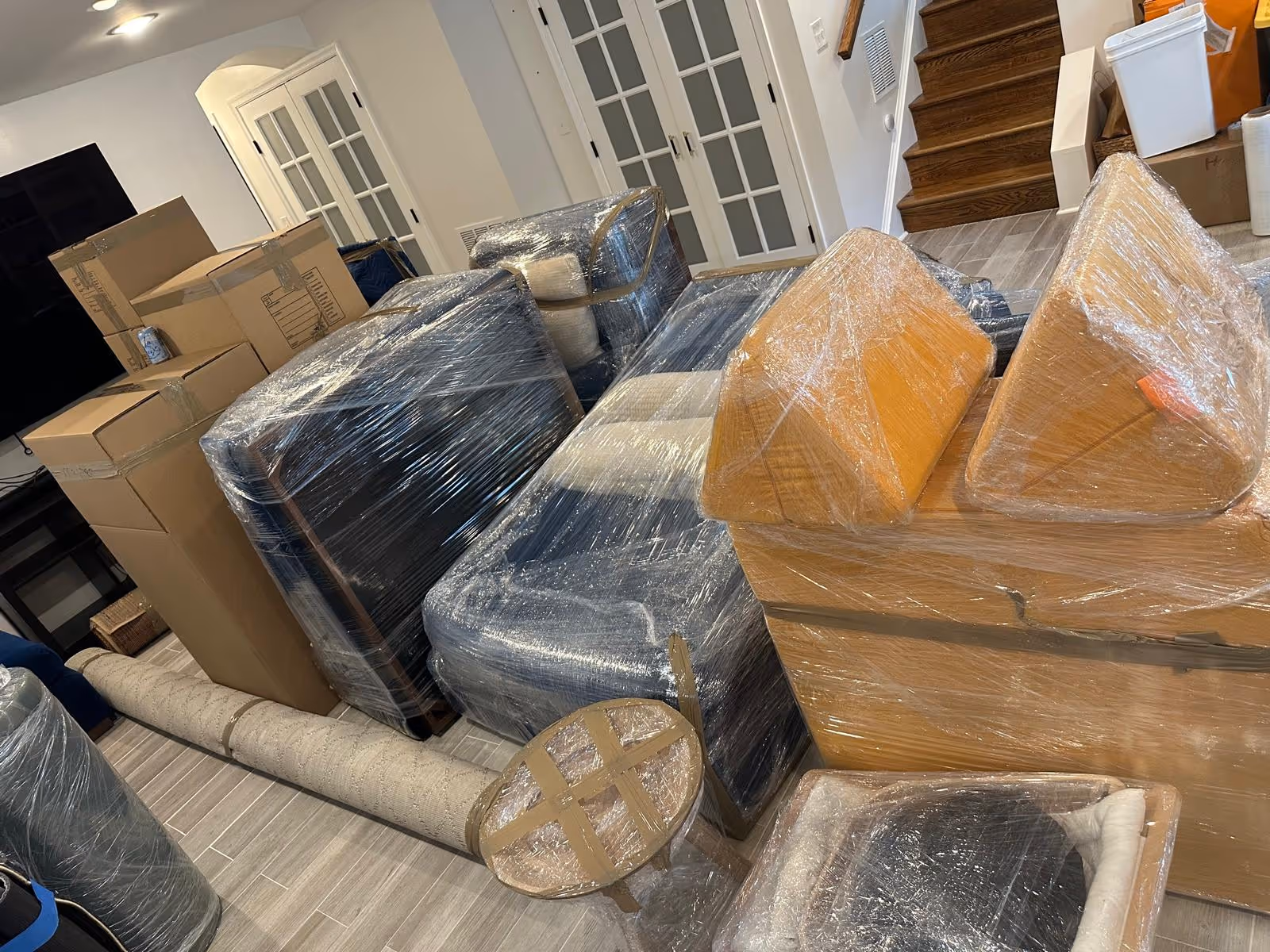 Several pieces of furniture and cardboard boxes wrapped in protective plastic and tape, placed indoors near stairs and French doors.
