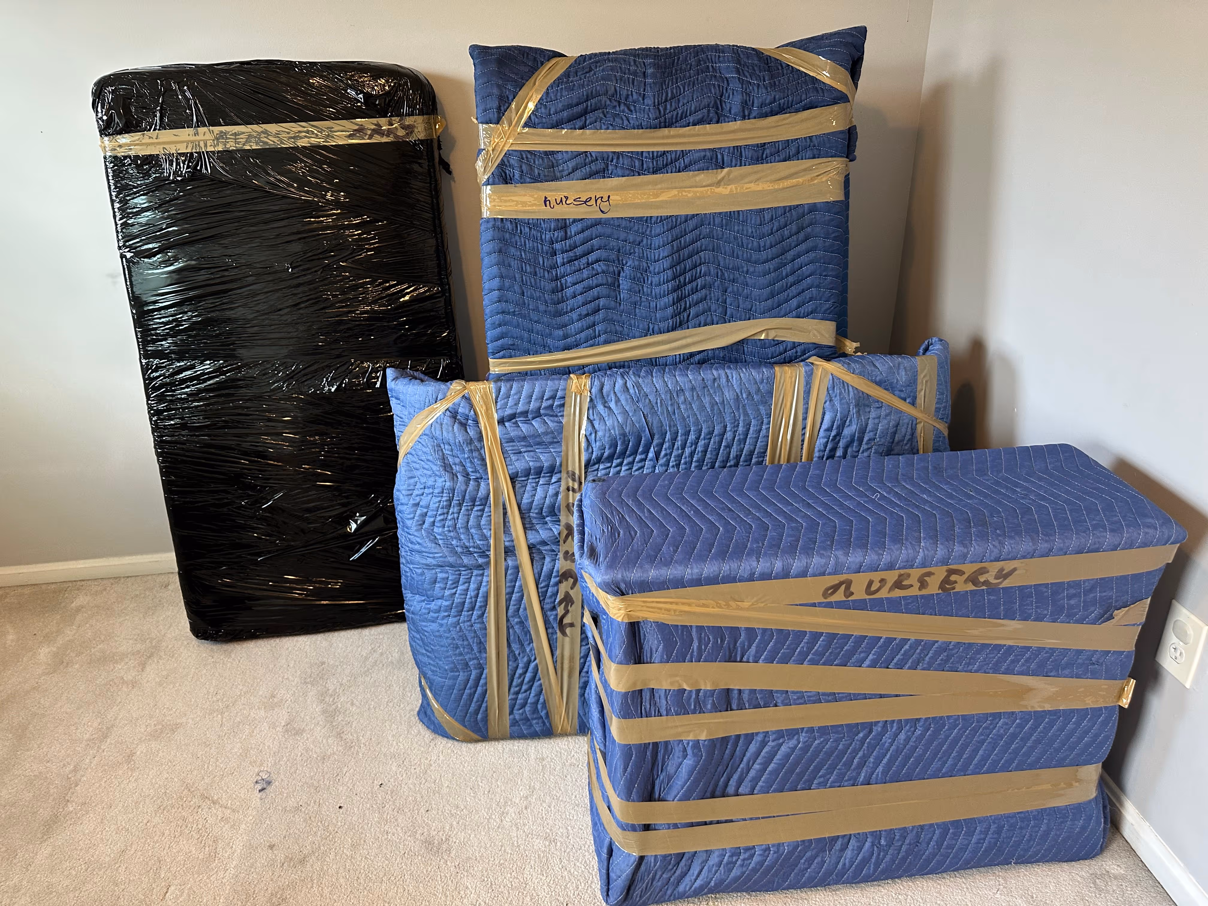 Four pieces of furniture wrapped in moving blankets and black plastic, secured with brown packing tape labeled 'nursery'.