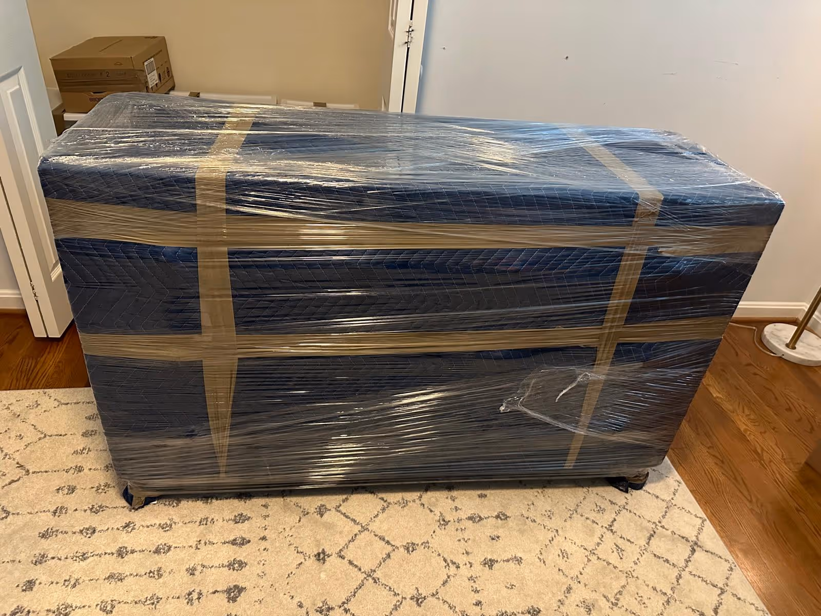 Large furniture piece wrapped in blue moving blankets and secured with tape and plastic wrap, placed on a patterned rug in a room.