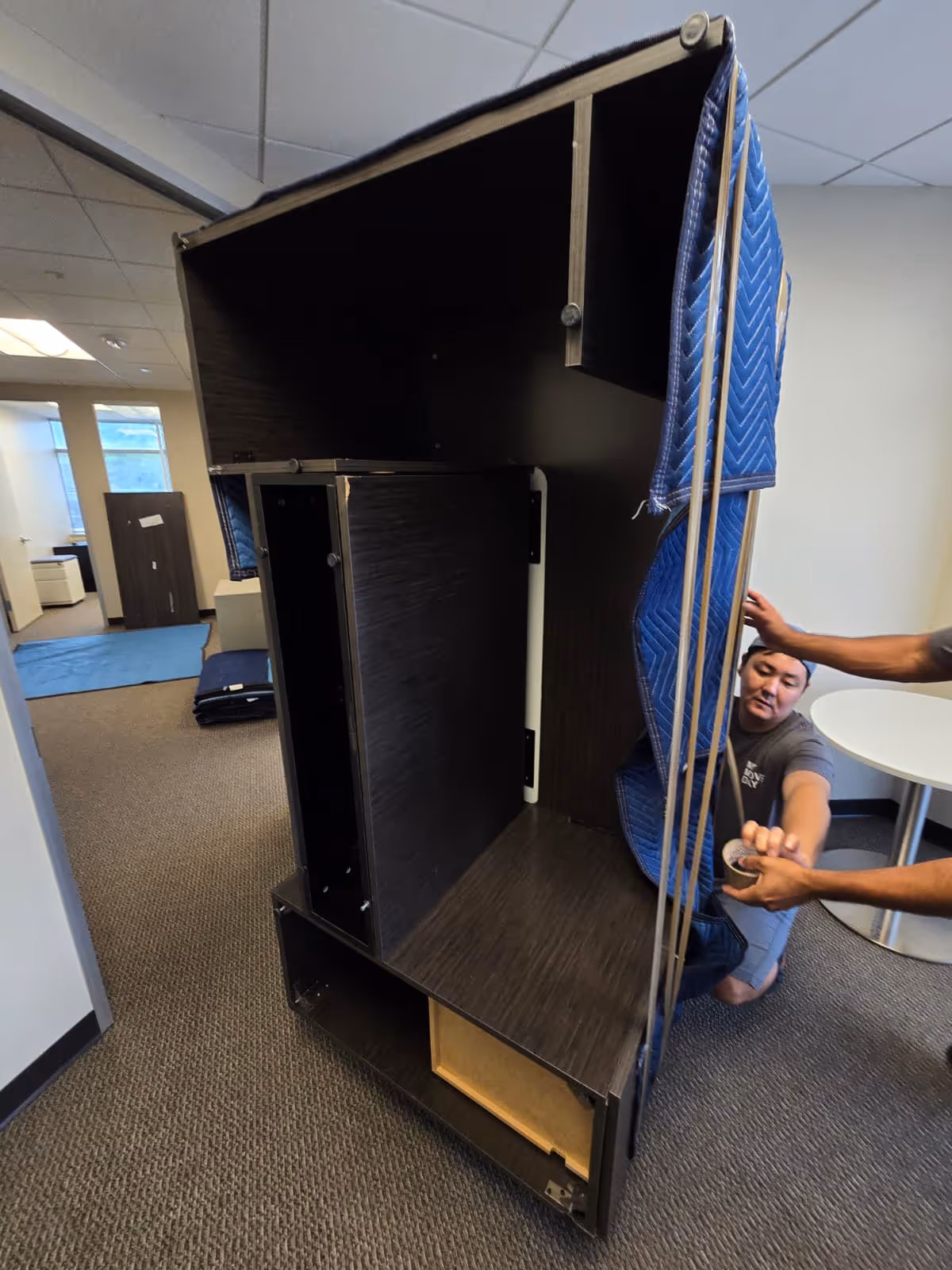 Two people wrapping and securing a large dark wood cabinet with a blue moving blanket and tape in an office space.