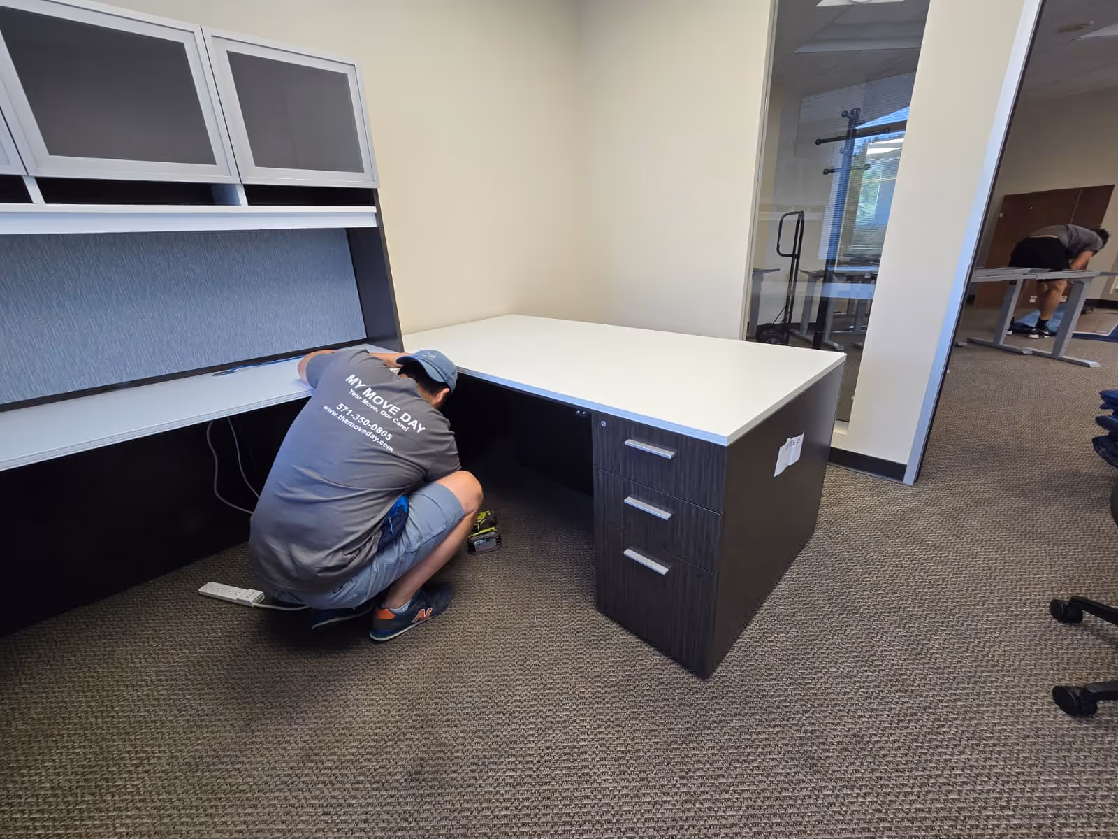 Two workers assembling or installing office desks in a carpeted room with beige walls and a glass partition.