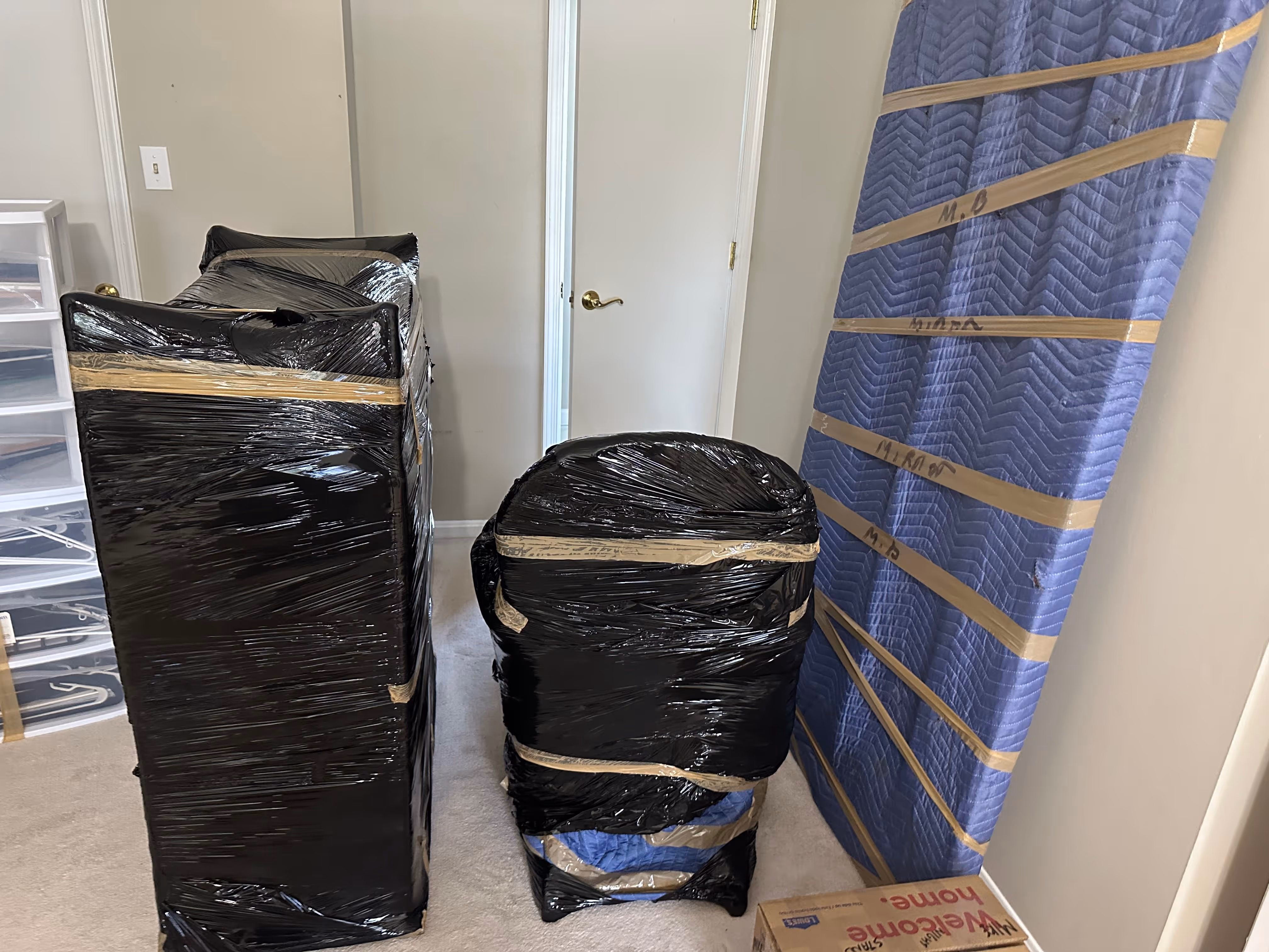 Three pieces of furniture wrapped in black plastic wrap and a blue moving blanket, standing upright in a room near a white door.