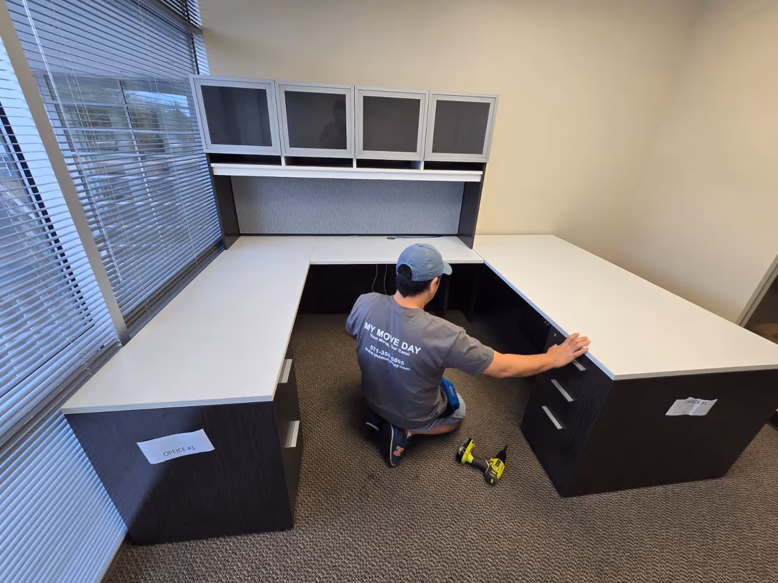 Man assembling or adjusting a U-shaped office desk with white surfaces and black drawers in a carpeted room.