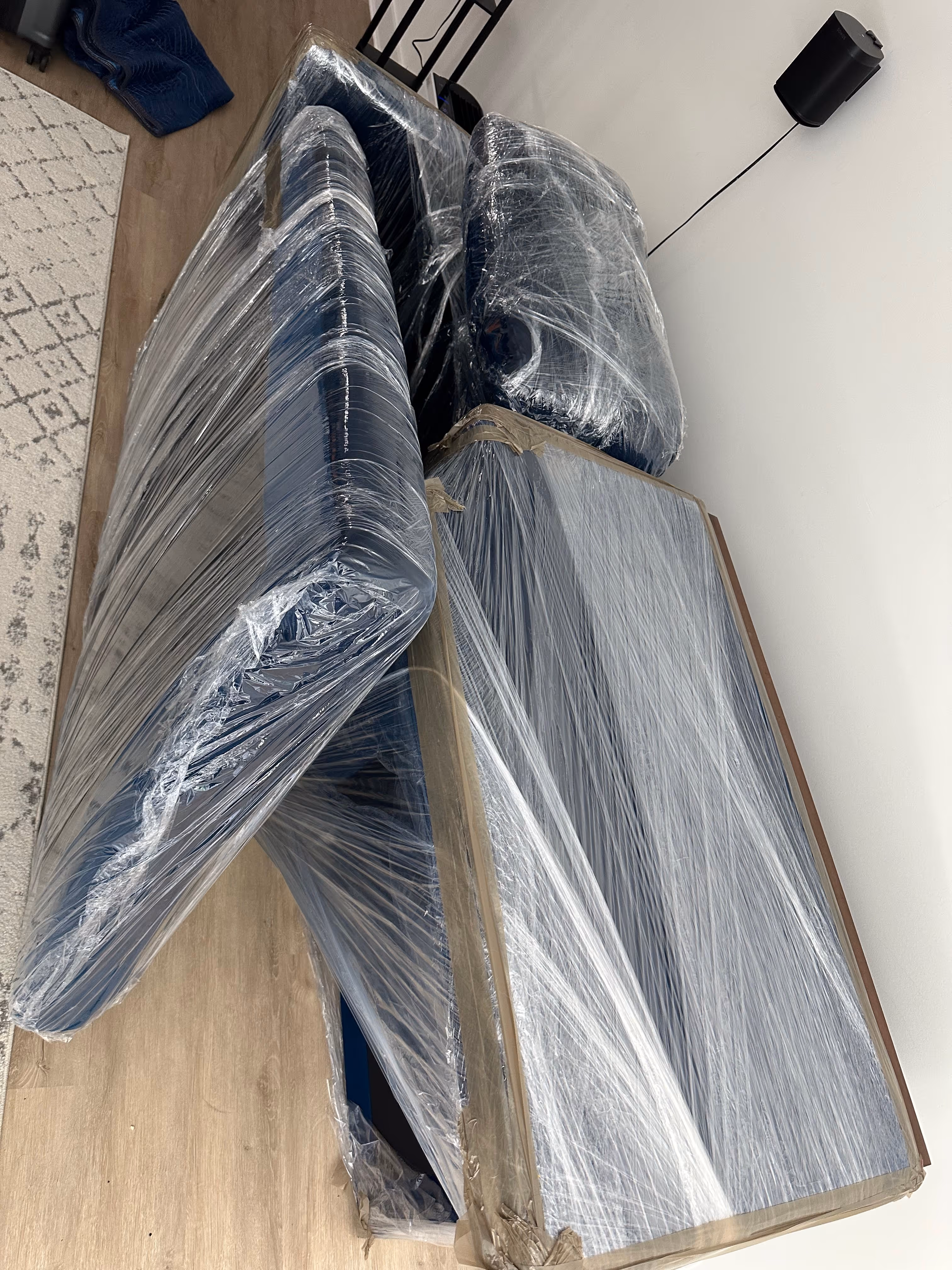 Furniture pieces wrapped tightly in clear plastic wrap placed on a wooden floor inside a room.