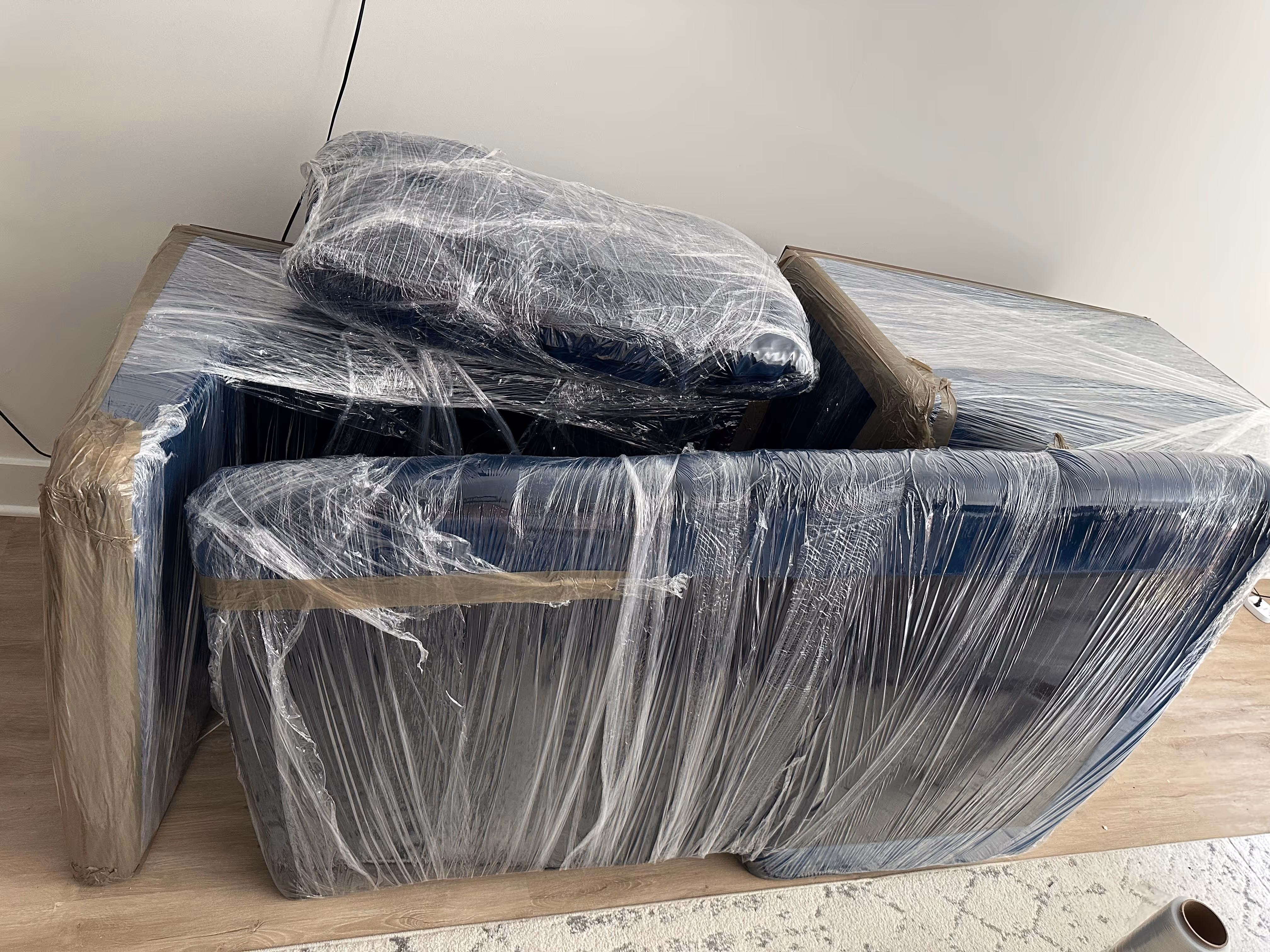 Blue furniture pieces wrapped securely in transparent plastic wrap on a wooden floor near a patterned rug.