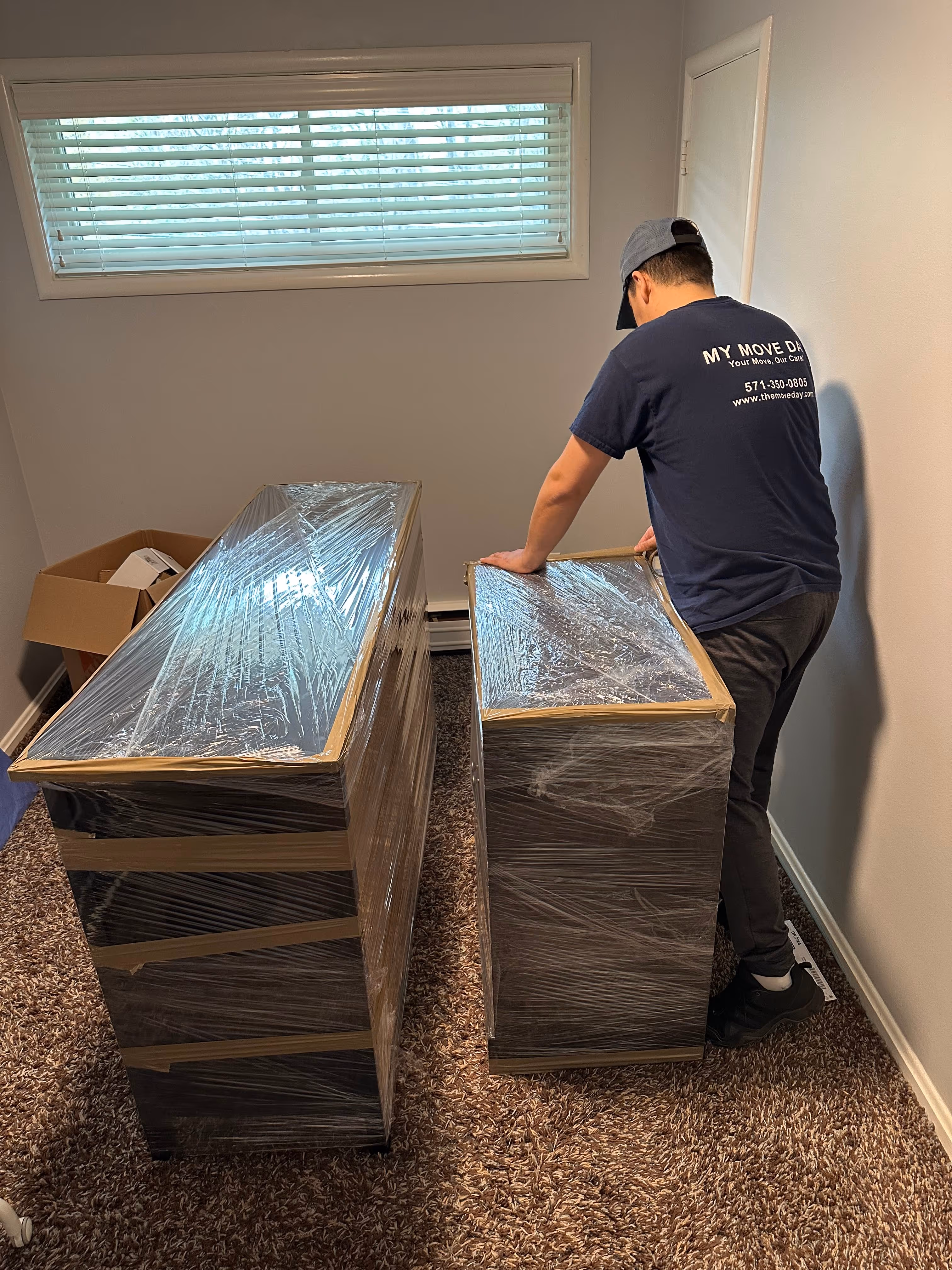 Man wearing a cap and black shoes wrapping furniture in plastic wrap inside a carpeted room with a window and a cardboard box.