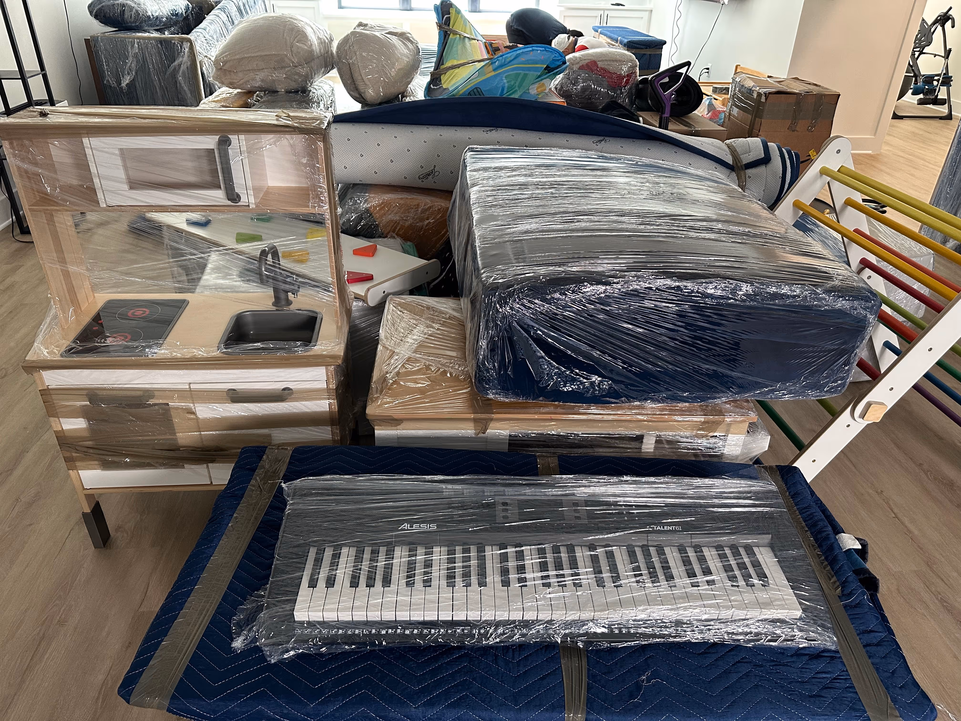 Various furniture and items wrapped in plastic and packing blankets, including a toy kitchen and an electronic keyboard, stacked indoors on a wooden floor.