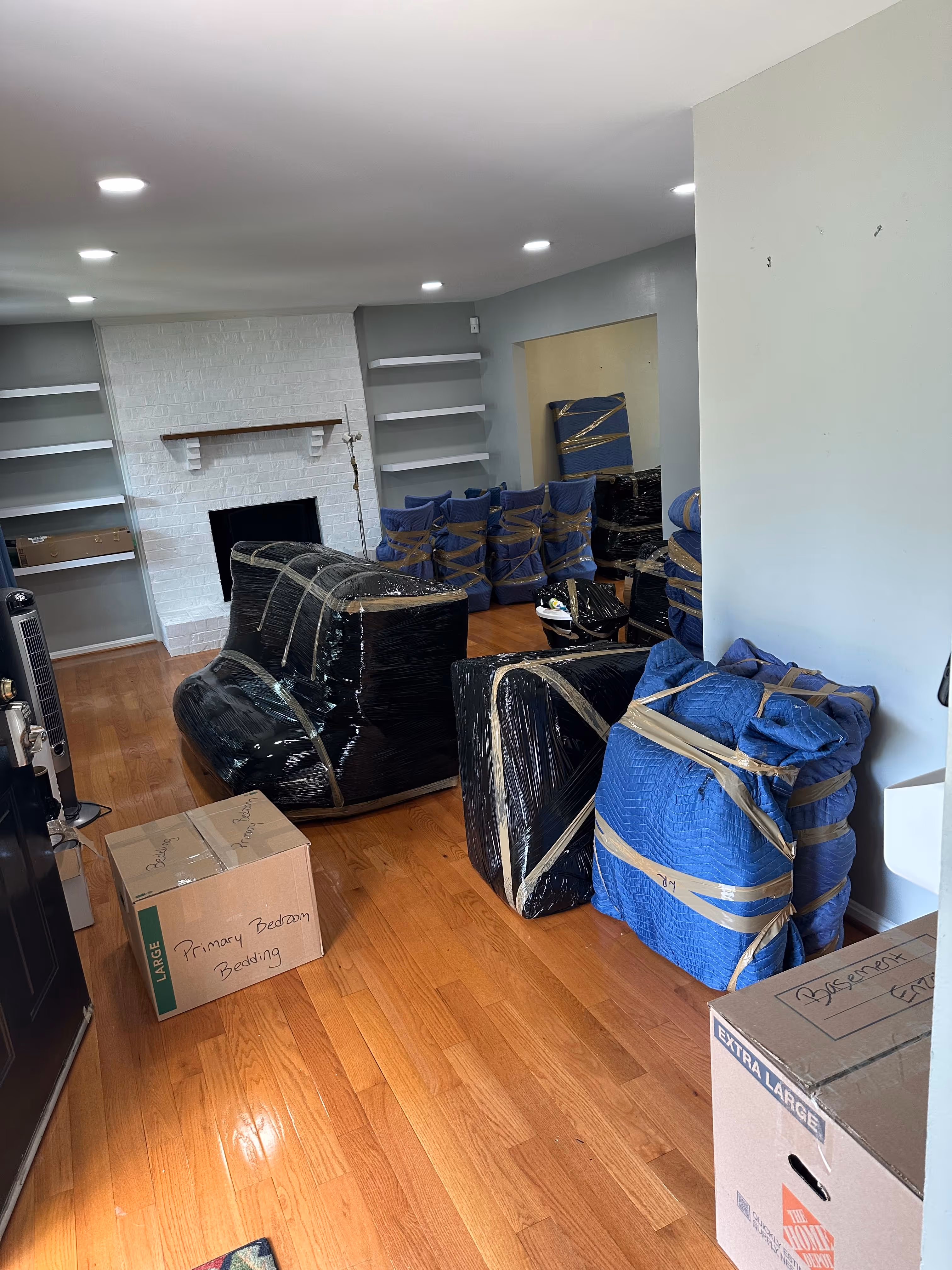 Living room with hardwood floor filled with furniture and boxes wrapped in protective covers and tape, ready for moving.