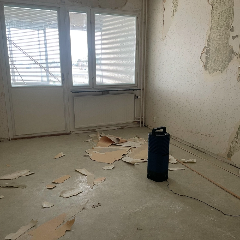 The Simulair device (a dark blue cylindrical unit) placed on the bare concrete floor of an empty apartment room mid-renovation, with stripped wallpaper debris scattered around it and windows with closed blinds in the background.