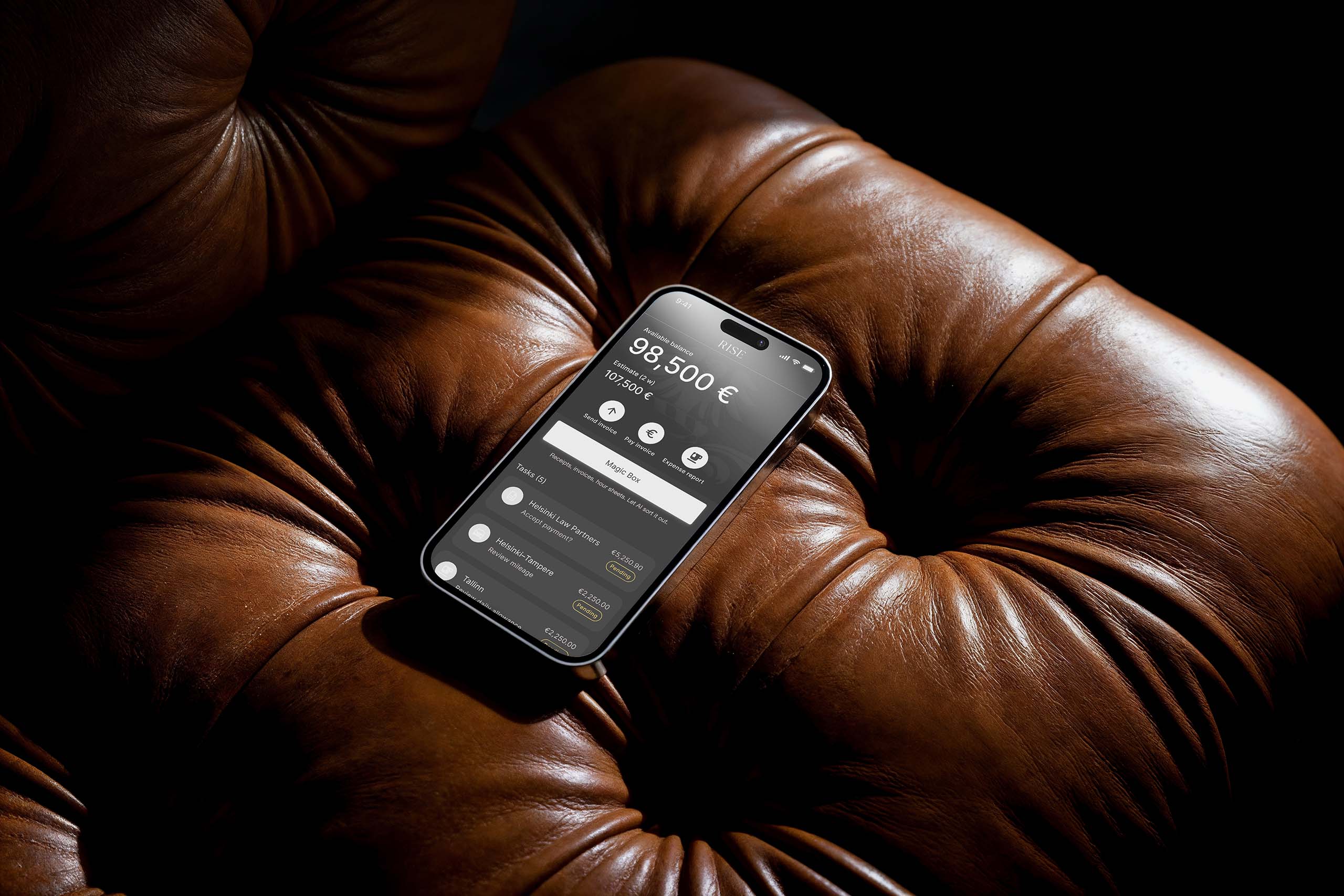 Smartphone displaying a financial app with balance and tasks interface on a brown leather cushioned surface.