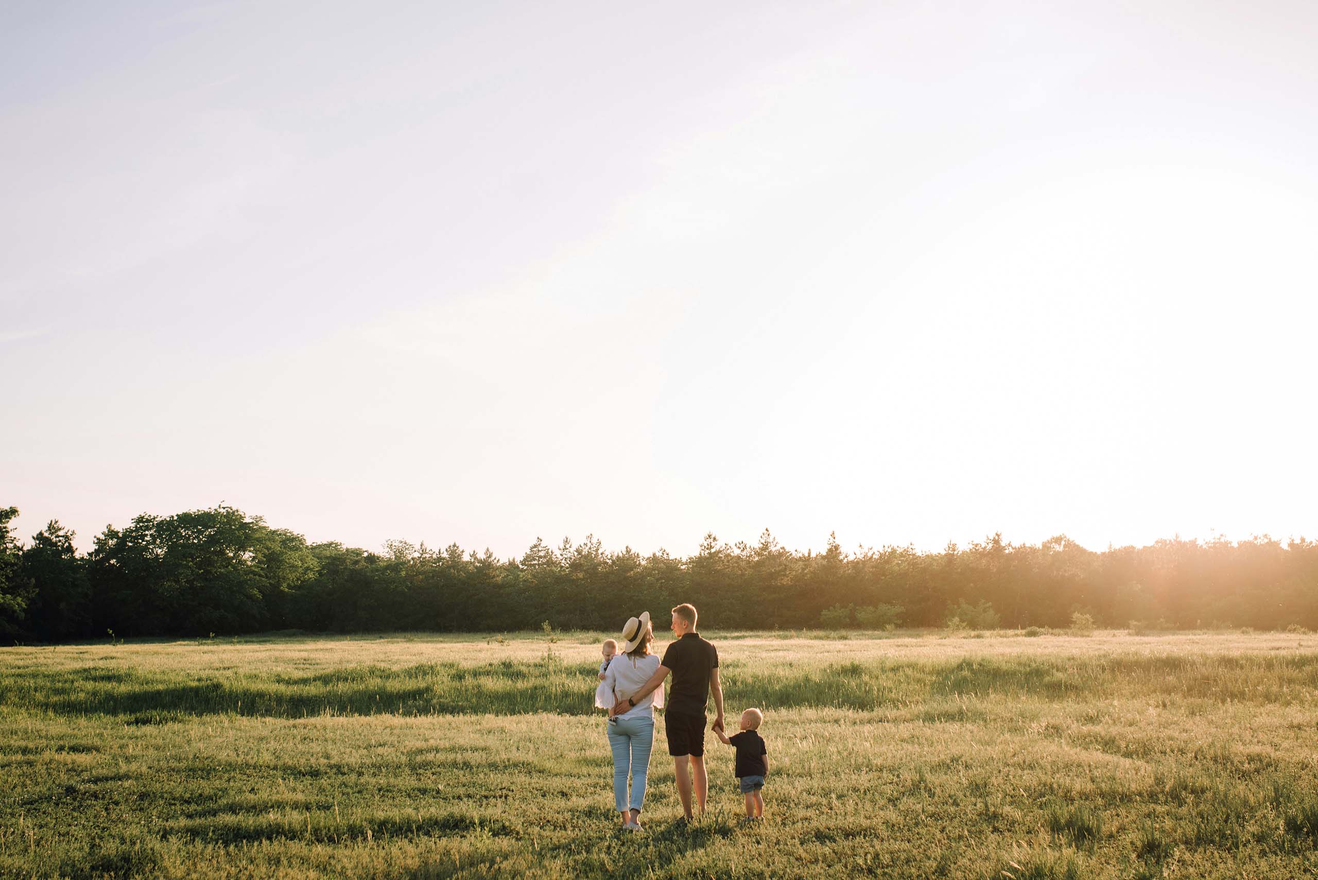 Family of four walking hand in hand through a sunlit grassy field toward a tree line at sunset.