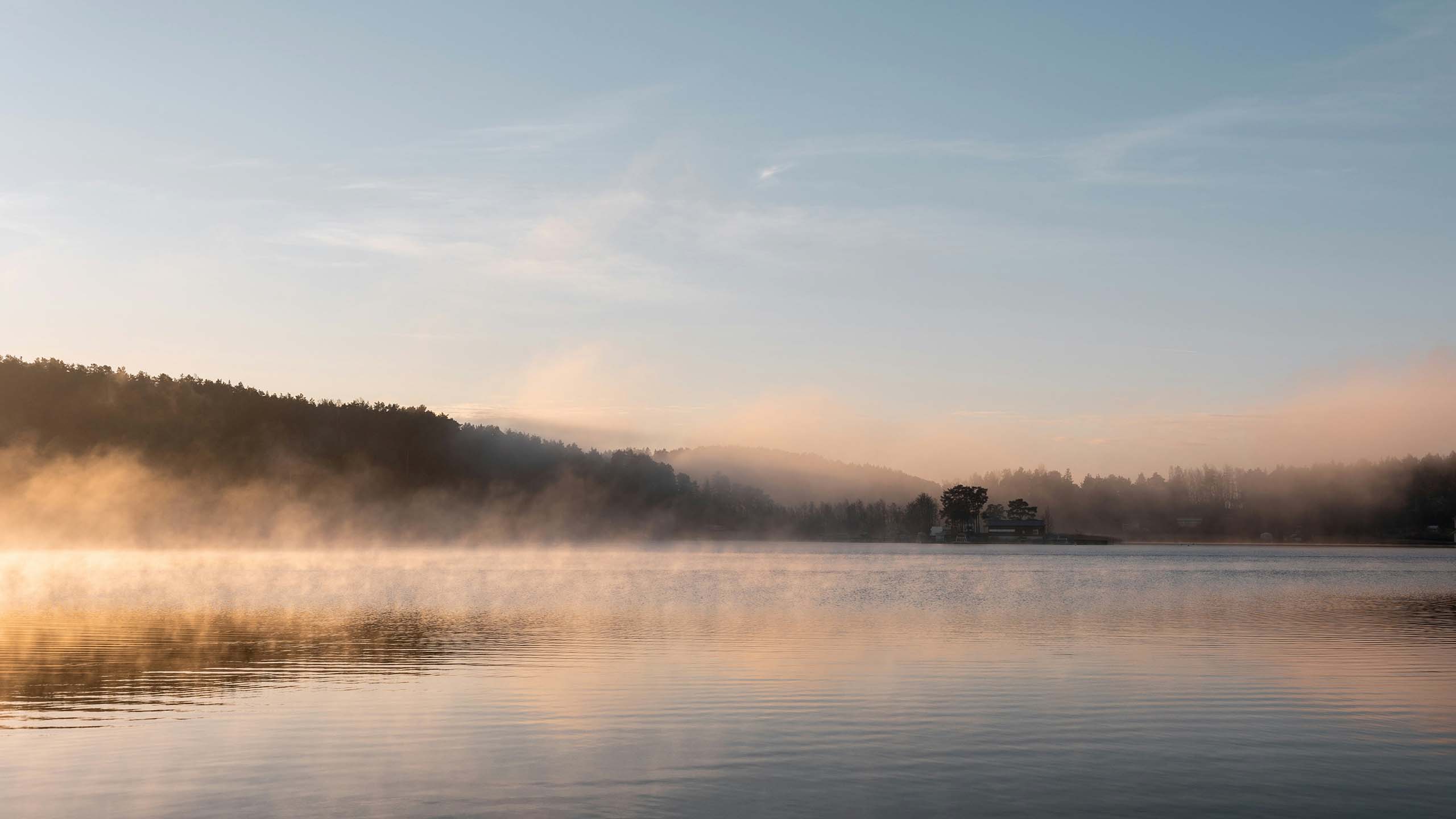 Calm lake with morning mist rising, surrounded by forested hills under a clear sky.