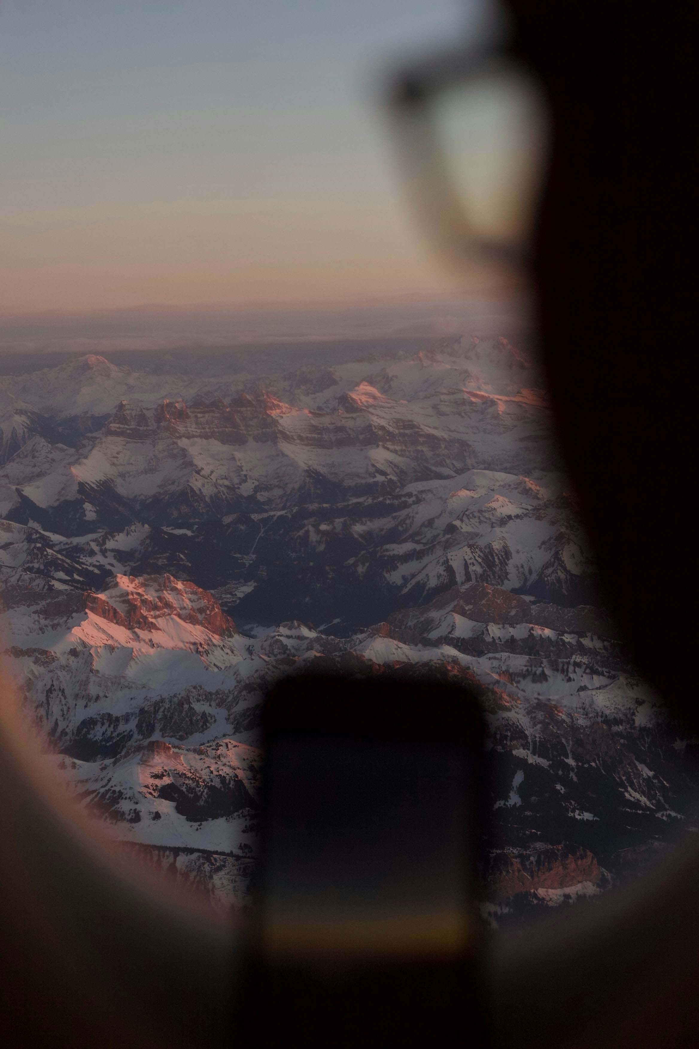 Snow-covered mountain peaks at sunset viewed from an airplane window with a phone silhouetted.