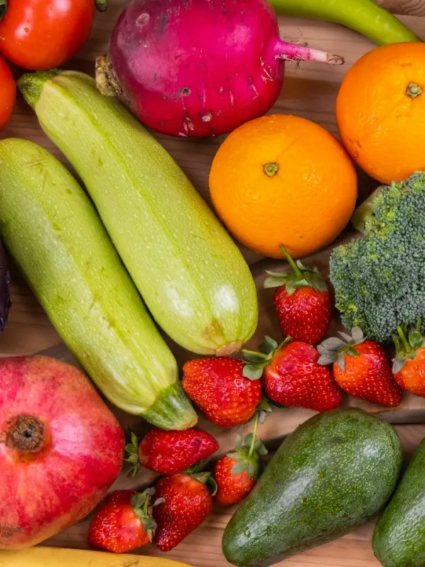 A variety of fresh fruits and vegetables including strawberries, oranges, zucchini, avocado, broccoli, pomegranate, and a red radish on a wooden surface.