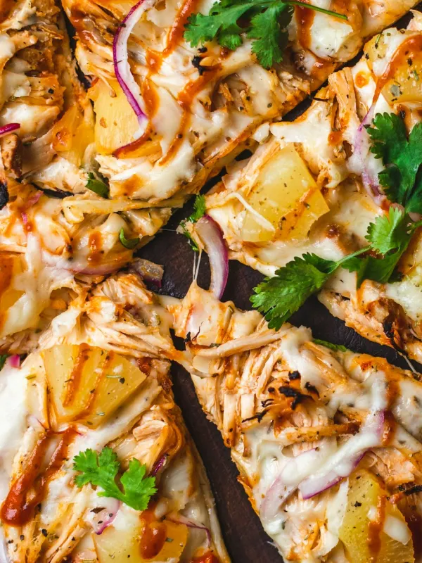 Close-up of barbecue chicken pizza slices topped with pineapple chunks, melted cheese, red onion slices, cilantro, and drizzled barbecue sauce.