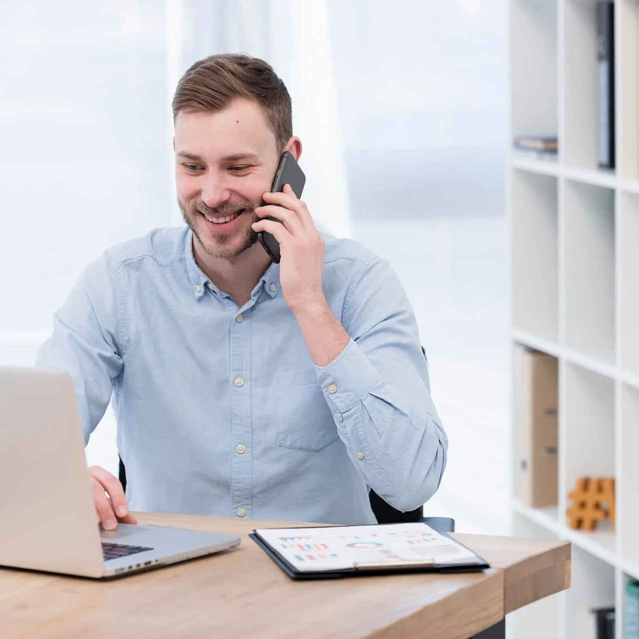 Smiling man in a light blue shirt talking on a smartphone while using a laptop at a wooden desk with a clipboard of charts.