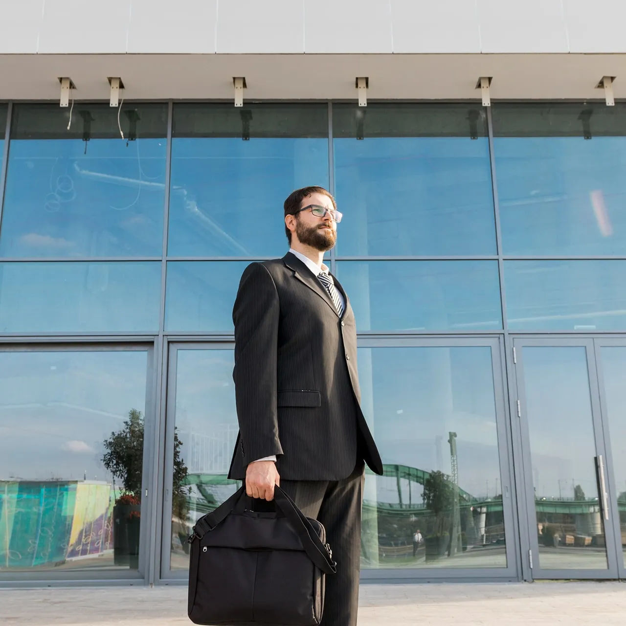 Man in a black suit and glasses holding a black briefcase standing in front of a large glass building.