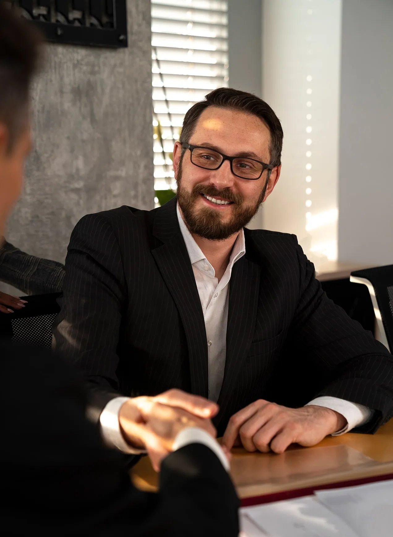 Smiling man with glasses and beard shaking hands across a table in an office setting.