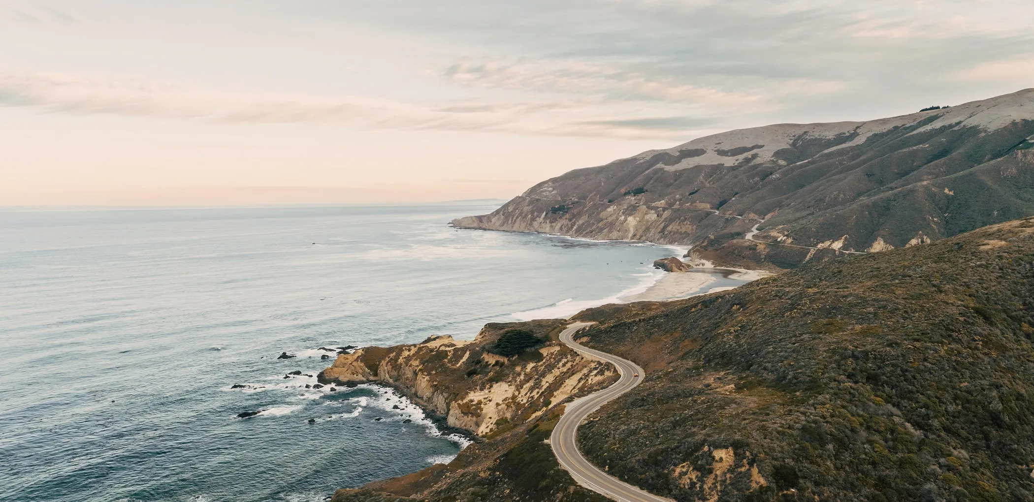 Curving coastal road along cliffs beside a calm ocean under a cloudy sky at sunset.
