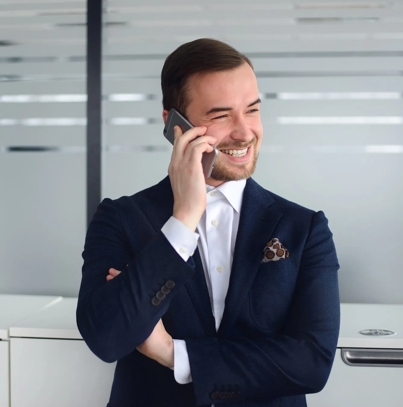 Smiling young man in a navy suit talking on a smartphone in an office setting.