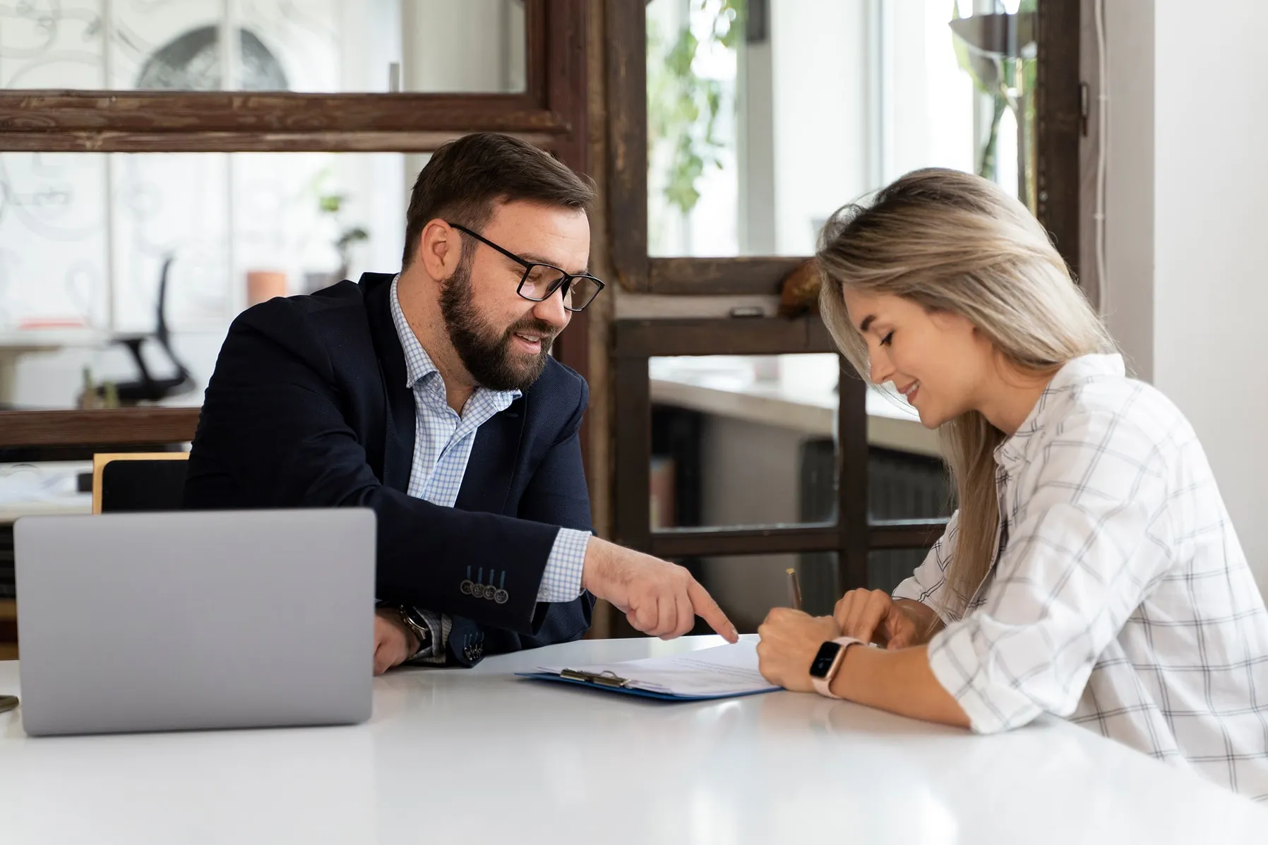 Smiling man in glasses pointing to a document while a woman in a checkered shirt signs it at a table with a laptop.