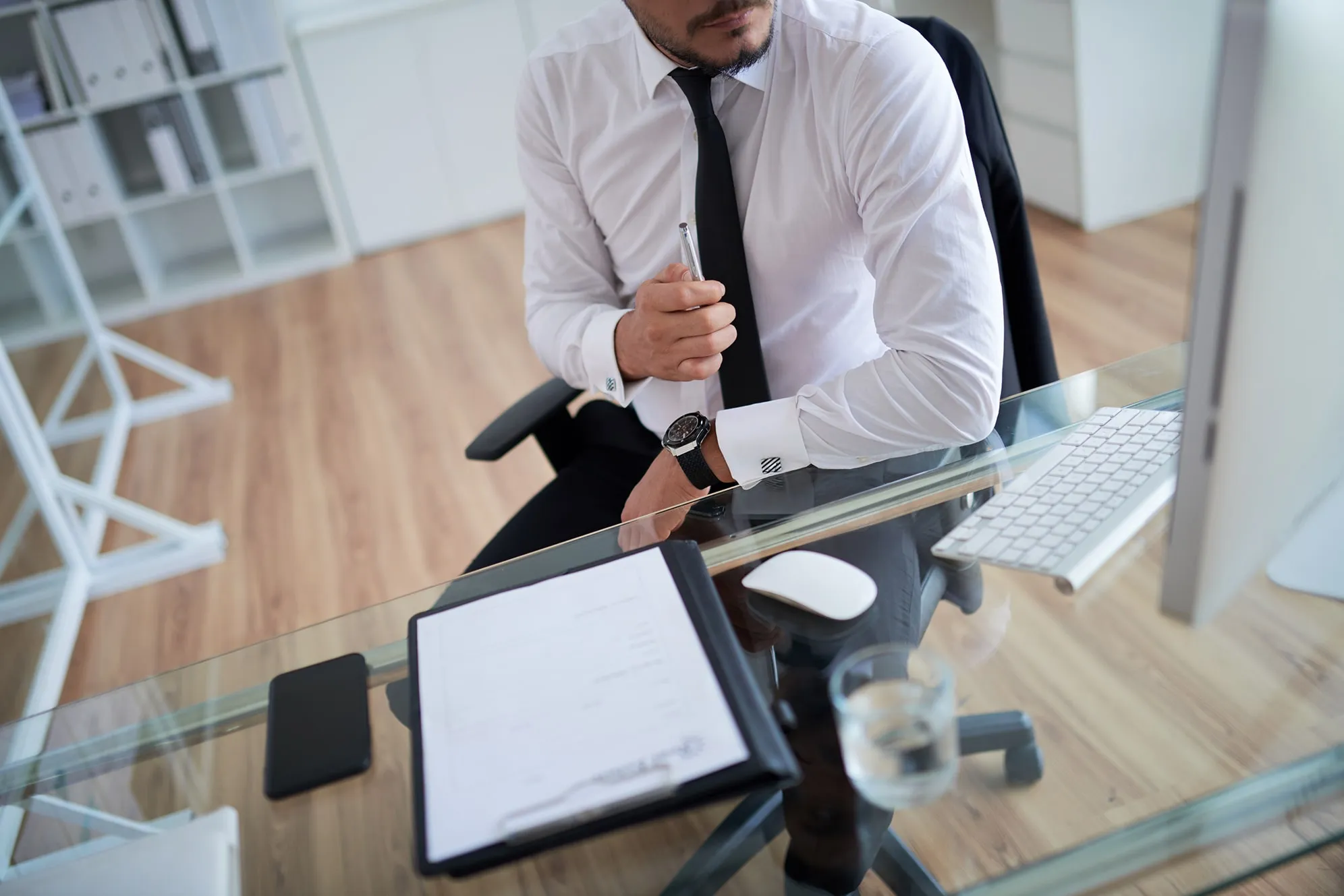 Man in white shirt and black tie sitting at a glass desk with keyboard, mouse, smartphone, clipboard, and glass of water.