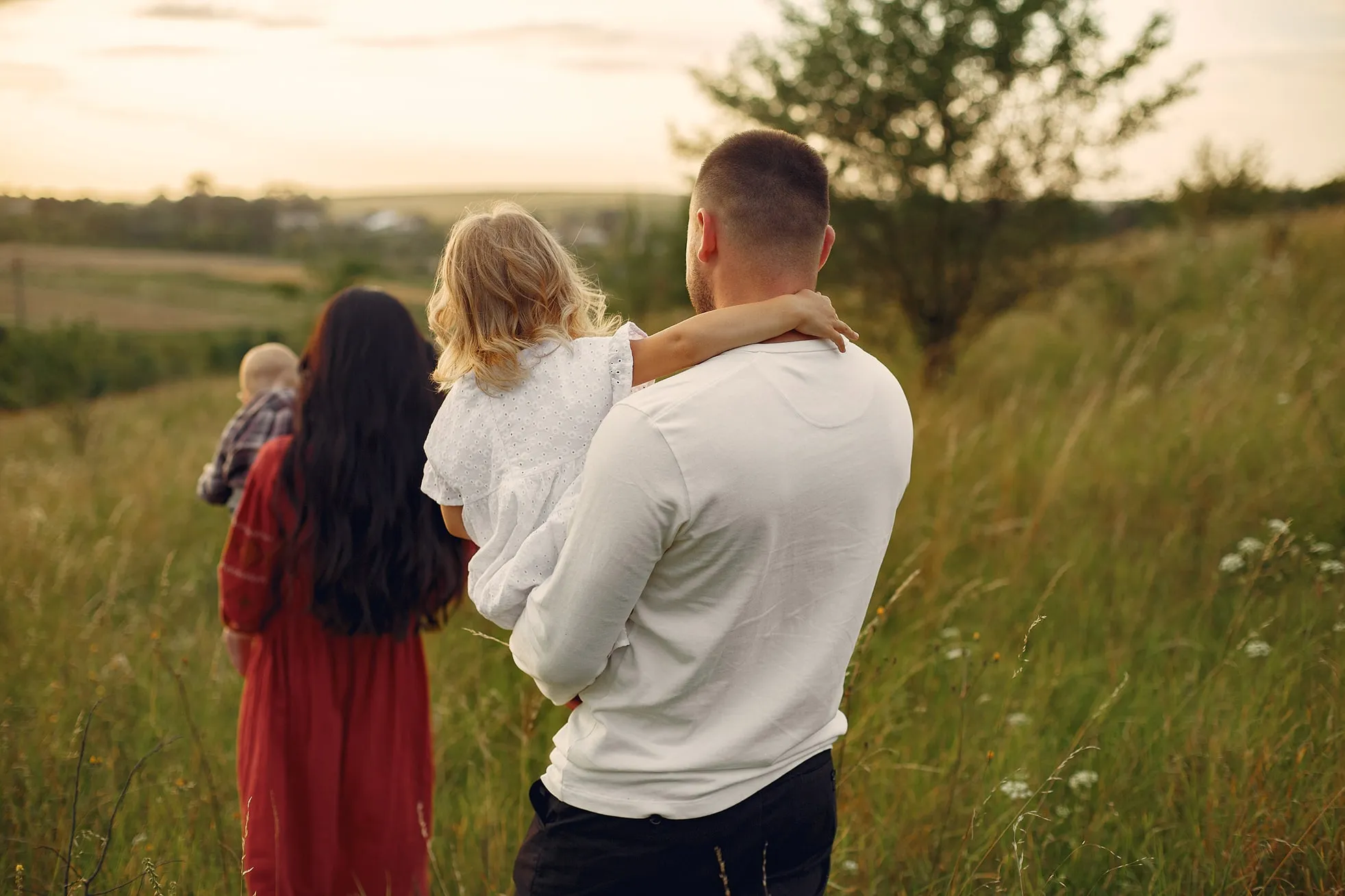 Man carrying young girl with arm around his neck and woman holding baby walking through a grassy field at sunset.
