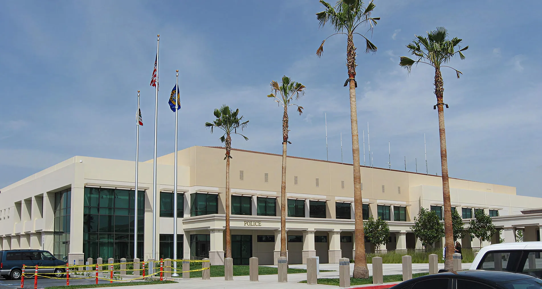 Modern police station building with several flagpoles and tall palm trees in front under a clear blue sky.