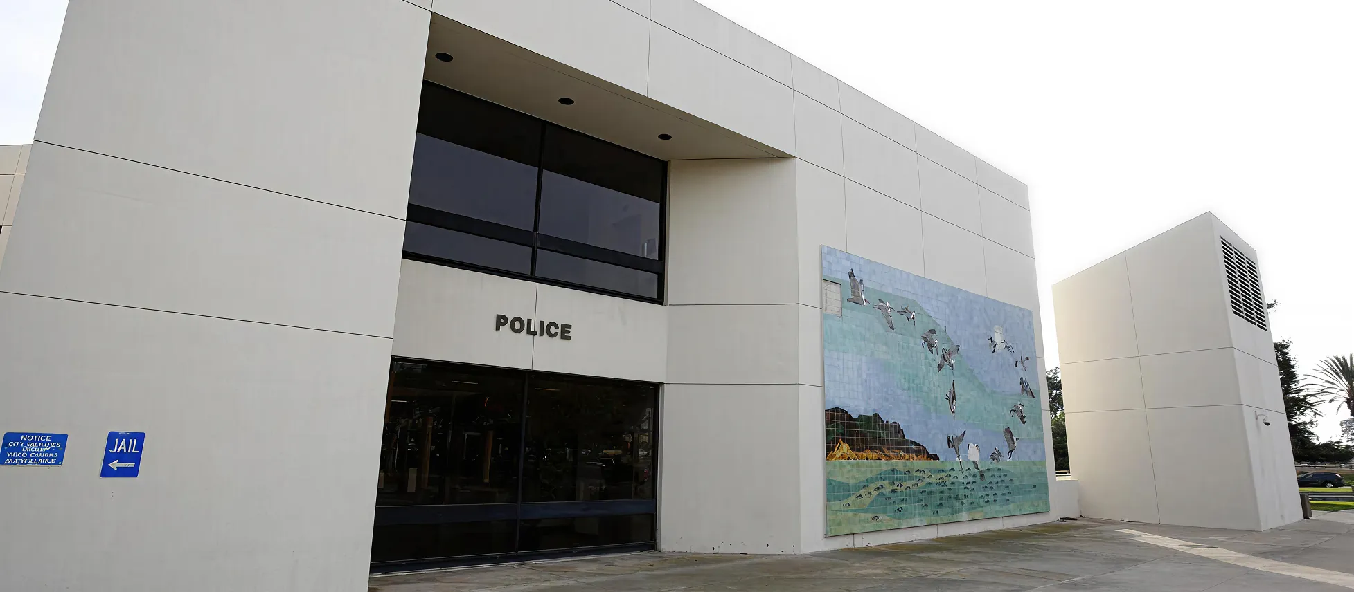 Exterior of a modern police station building with a large tiled mural of flying birds on the wall and blue signs indicating the direction to the jail.