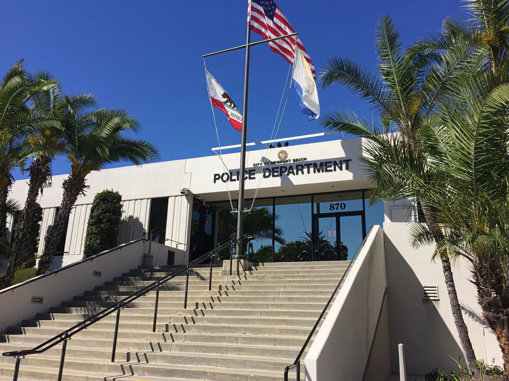 Entrance of Newport Beach Police Department building with stairs, palm trees, and three flags on flagpoles against a clear blue sky.