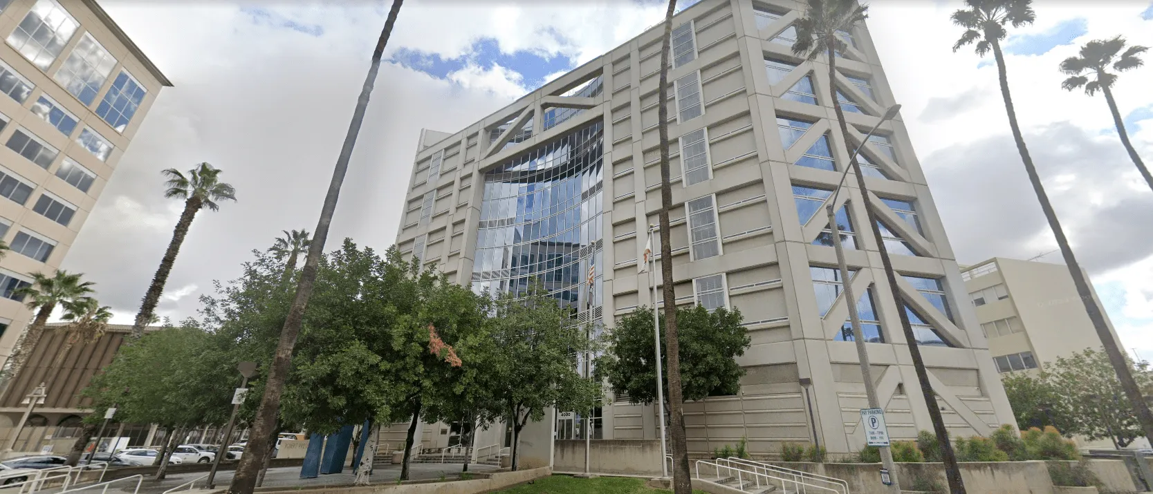 Modern multi-story office building with a glass facade and surrounding palm trees under a partly cloudy sky.