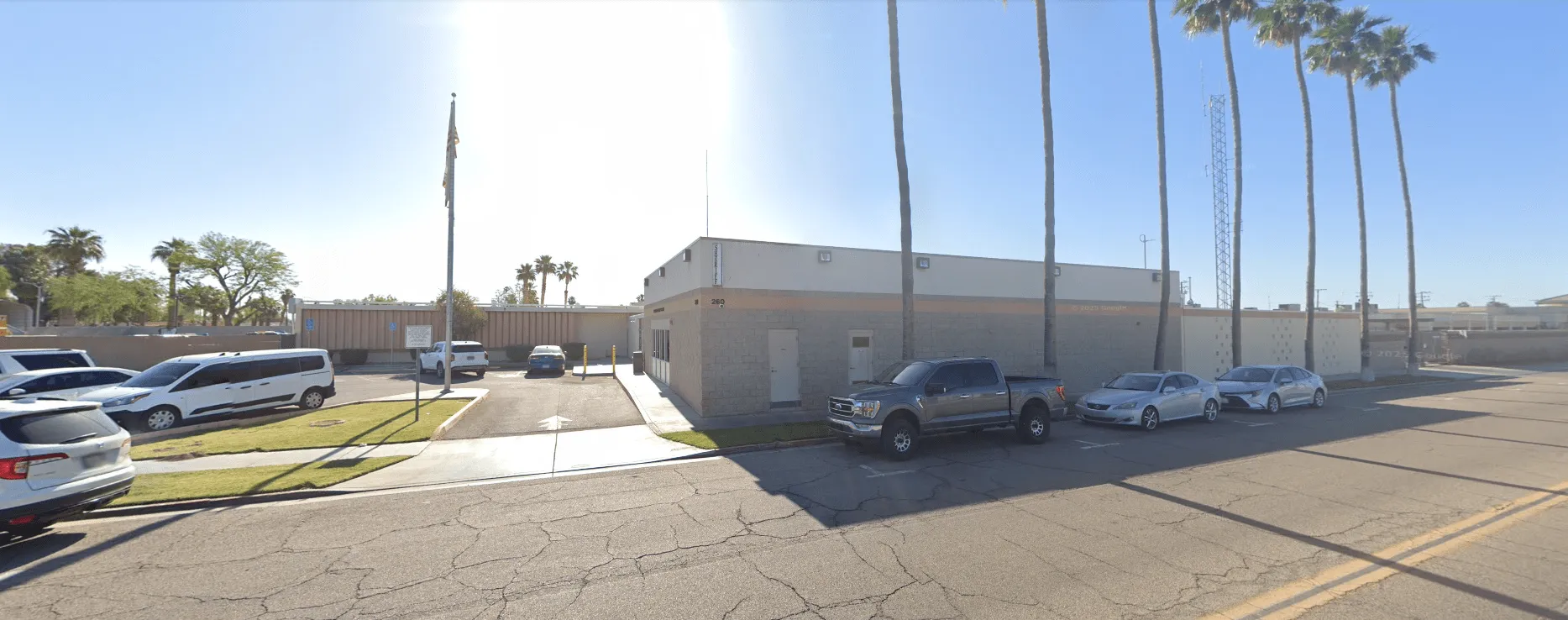 Single-story beige building with several parked cars and tall palm trees under a clear blue sky.