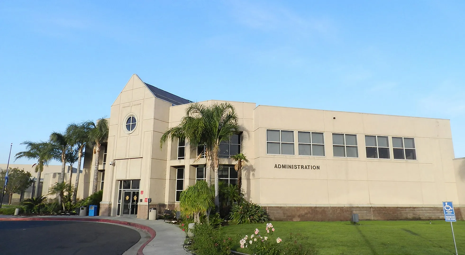 Two-story beige administration building with palm trees and landscaping in front under a clear blue sky.