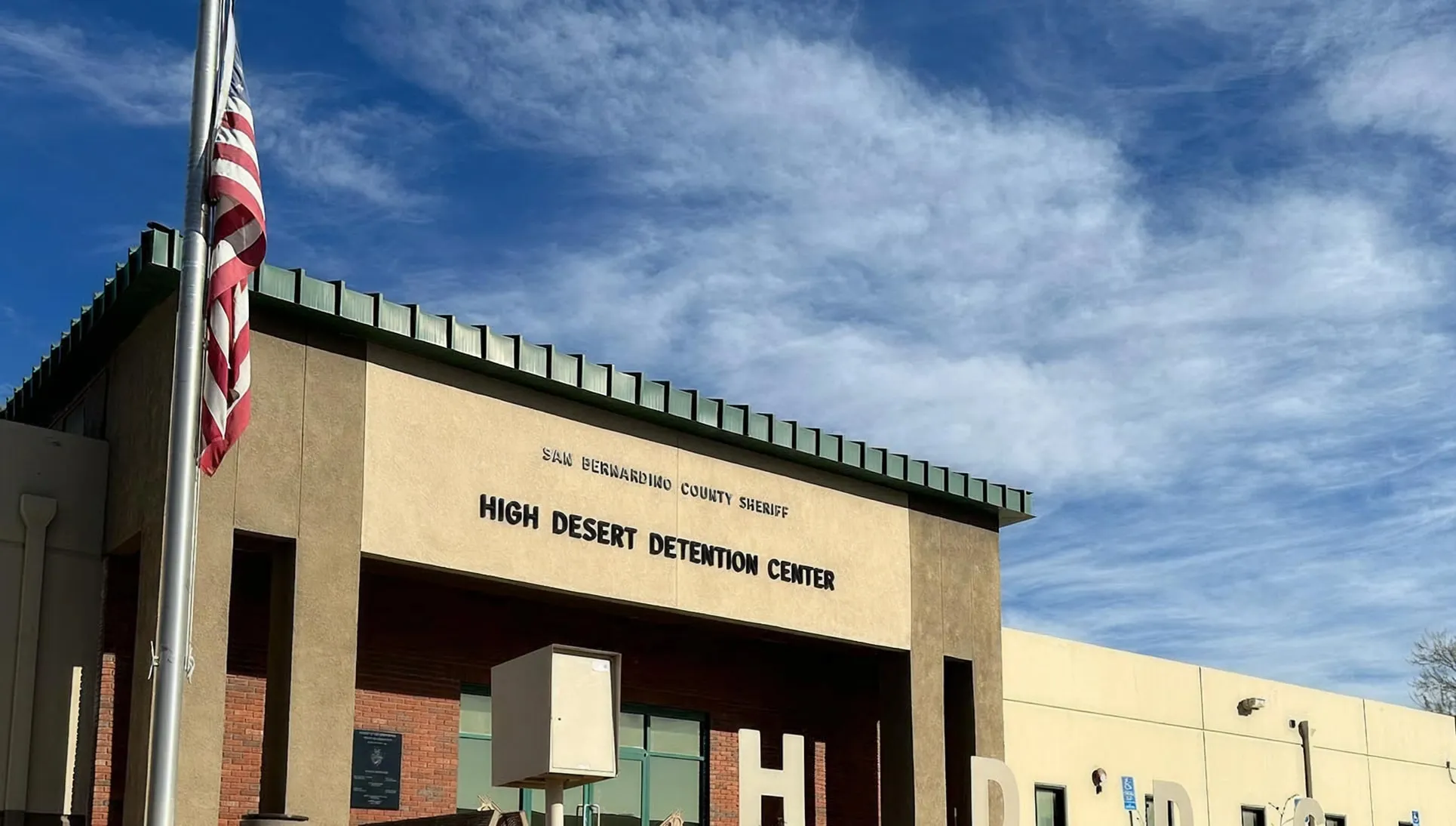 Exterior of the High Desert Detention Center building under a partly cloudy blue sky with a folded American flag on a flagpole in front.