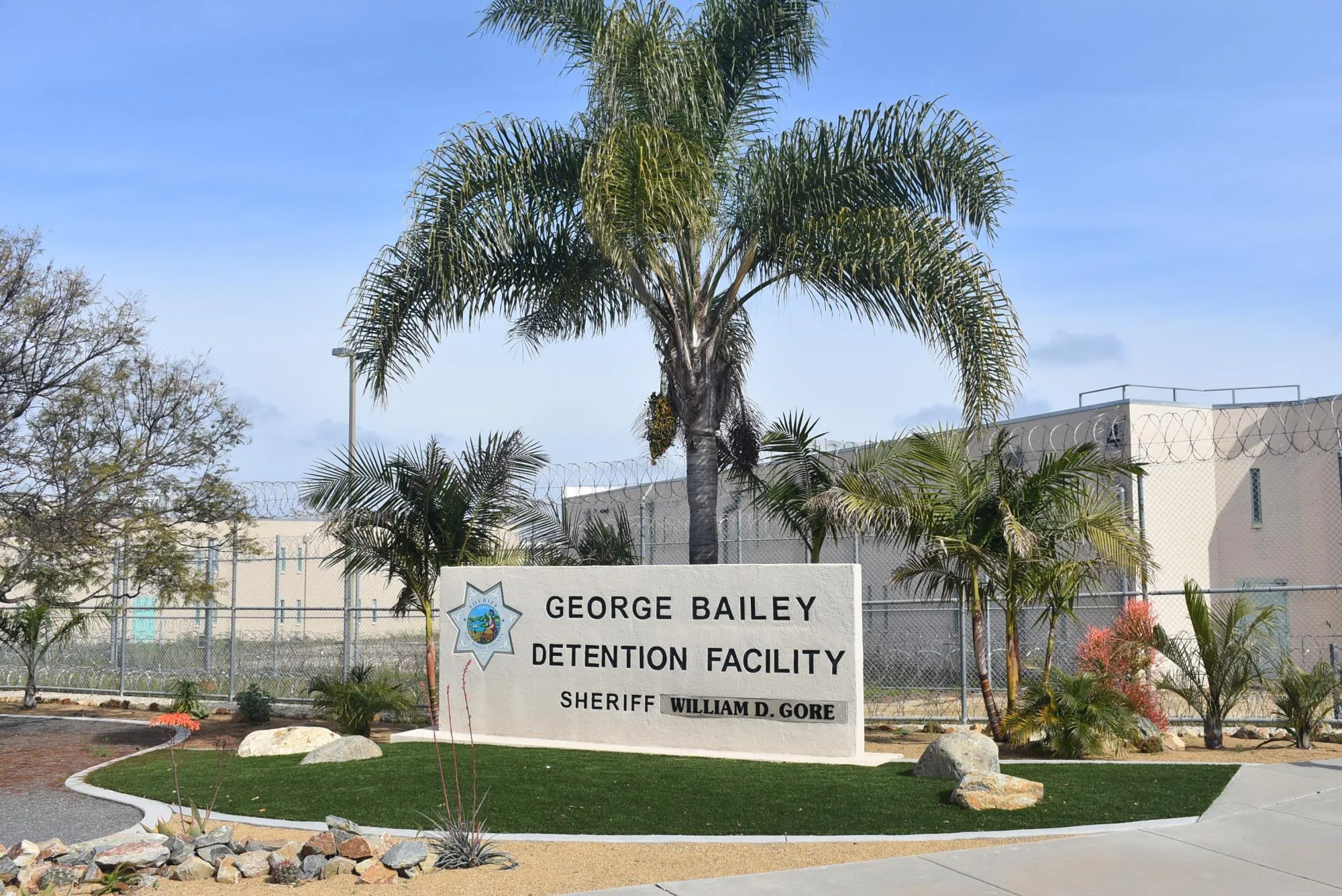 Entrance sign for George Bailey Detention Facility with surrounding palm trees and a chain-link fence topped with barbed wire.