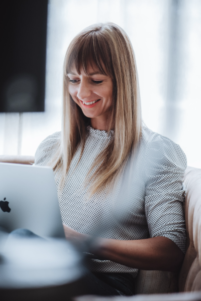 Auf dem Bild ist die Gründerin des Nomad Graphic zu sehen. Sie hat blonde lange Haare, lächelt nett und trägt eine weiße Bluse. Sie sitzt auf einer Couch und arbeitet am Laptop.