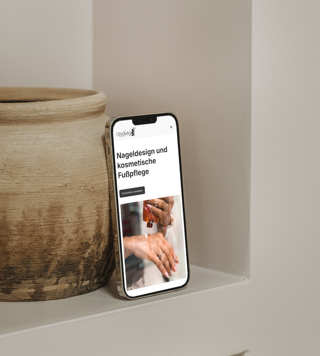 Ein iPhone, auf dem die Website des Ma Jolie Beauty Studios zu sehen ist. Die Seite zeigt ein elegantes Design mit einem großen Header-Bild von einer Hand einer Frau und bietet Informationen über Nageldesign und kosmetische Fußpflege.