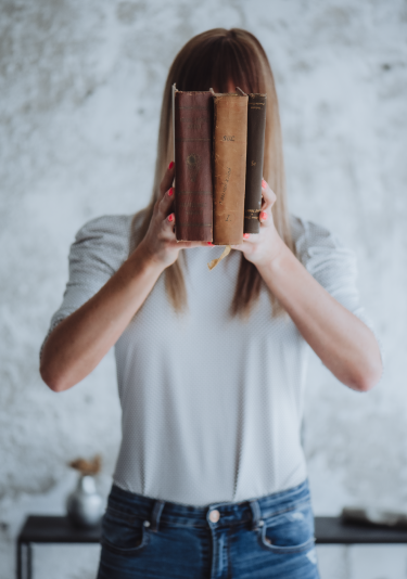 Auf dem Bild ist die Gründerin von Nomad Graphic zu sehen, sie hält drei Bücher vor ihr Gesicht. Sie trägt eine weiße Bluse und Jeans. Der Hintergrund ist eine helle, strukturierte Wand.