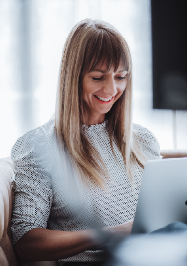 Auf dem Bild ist die Gründerin des Nomad Graphic zu sehen. Sie hat blonde lange Haare, lächelt nett und trägt eine weiße Bluse. Sie sitzt auf einer Couch und arbeitet am Laptop.