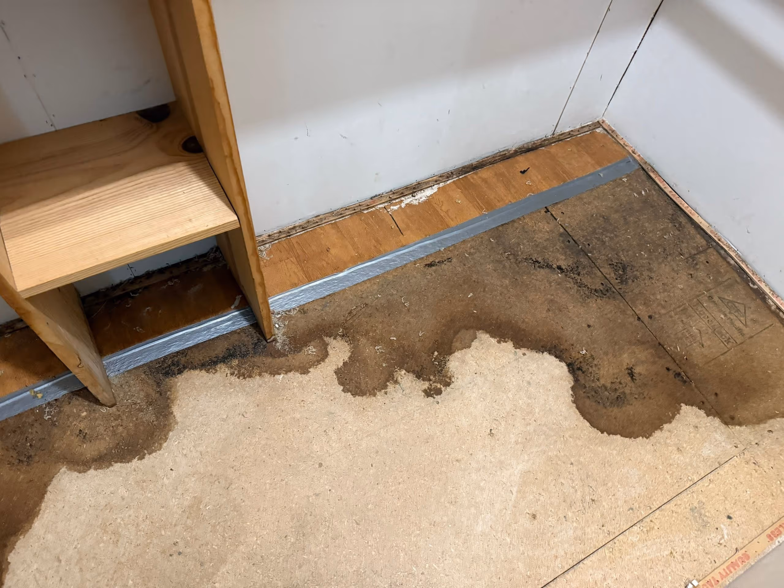 Corner of a room with wooden shelves and water-damaged plywood floor showing dark stains along the edges.